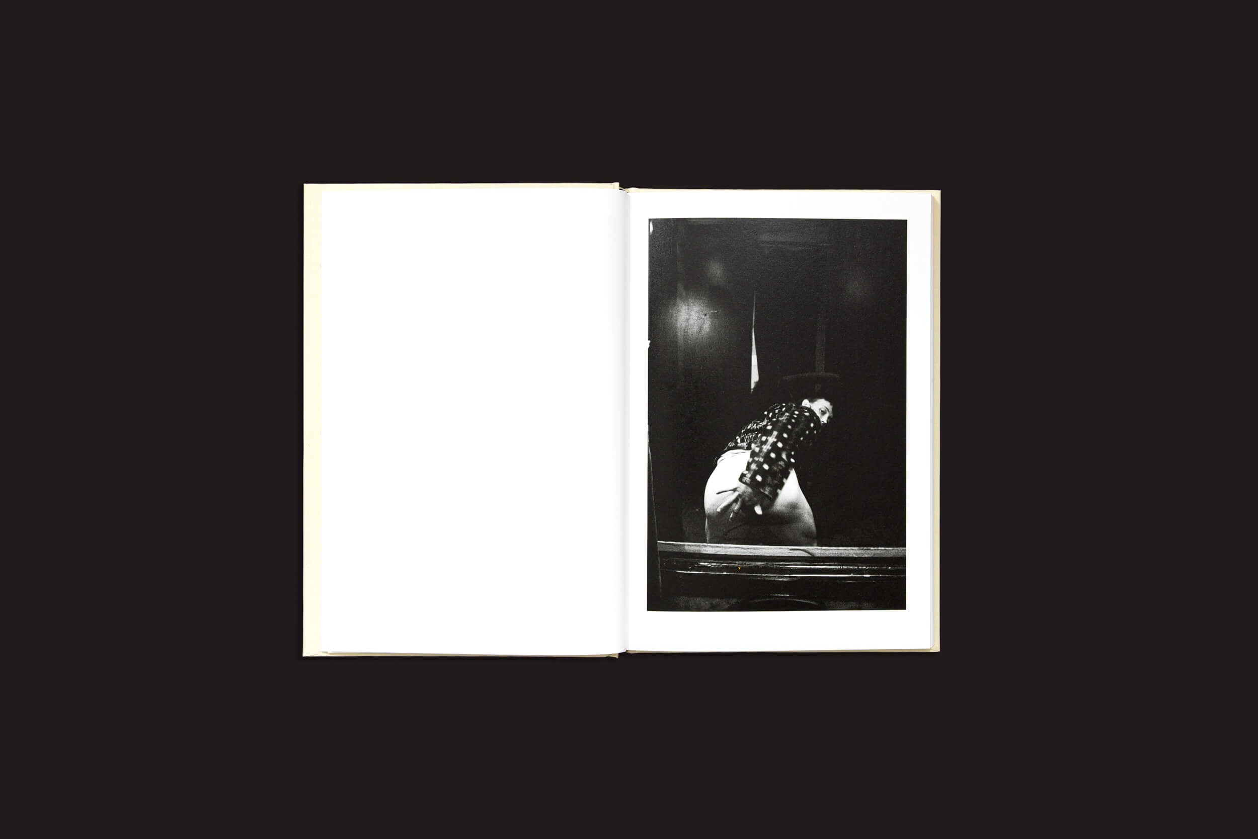 Um exemplar de "Cammie Toloui: 5 Dólares por 3 Minutos", de Void, está aberto sobre um fundo escuro. A página da esquerda está em branco, e a da direita exibe uma foto em preto e branco retratando a sexualidade masculina com alguém debruçado sobre uma superfície reflexiva, vestindo uma camisa estampada.