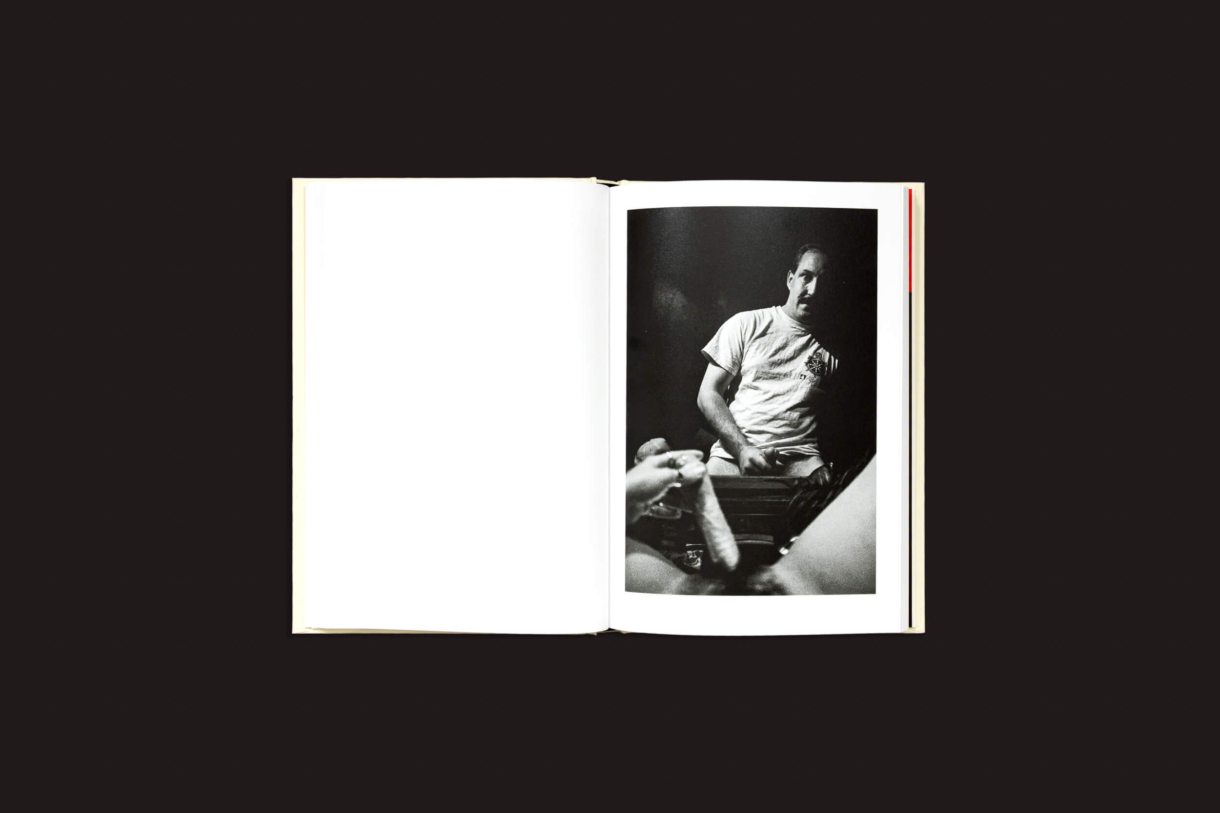 Em "Cammie Toloui: 5 Dólares por 3 Minutos", de Void, um livro aberto apresenta uma foto em preto e branco na página direita, retratando uma pessoa debruçada sobre uma mesa, vestindo uma camiseta lisa, explorando a sexualidade masculina. A página esquerda está em branco contra o fundo escuro.