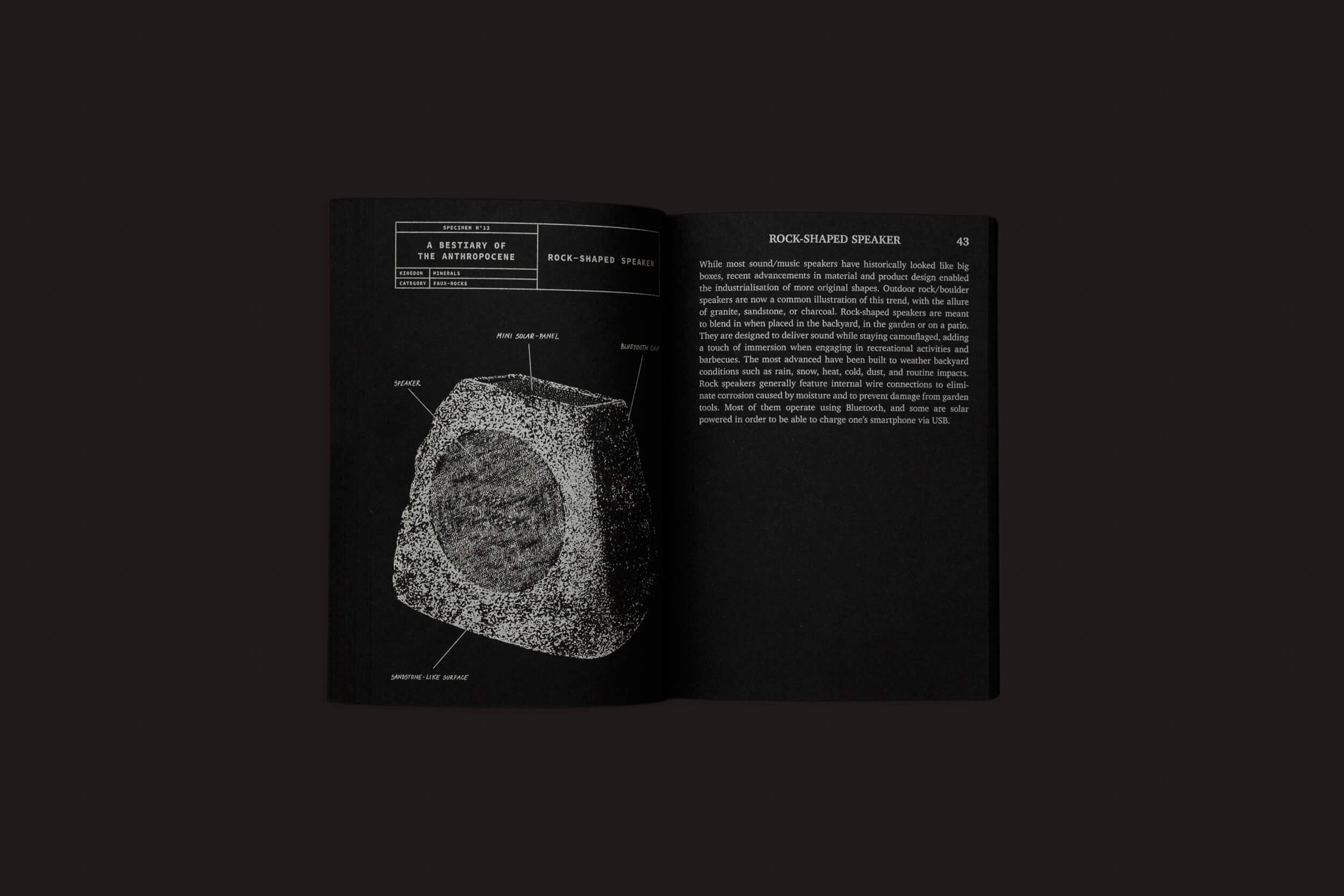 Um livro aberto repousa sobre uma superfície escura. A página da esquerda mostra um diagrama de rocha com rótulos; a da direita apresenta um texto intitulado "Rock-Shaped Speaker". Esta edição, "A Bestiary of the Anthropocene" de Nicolas Nova & Disnovation.org da Set Margins', captura uma era pós-natural com páginas pretas e impressão branca.