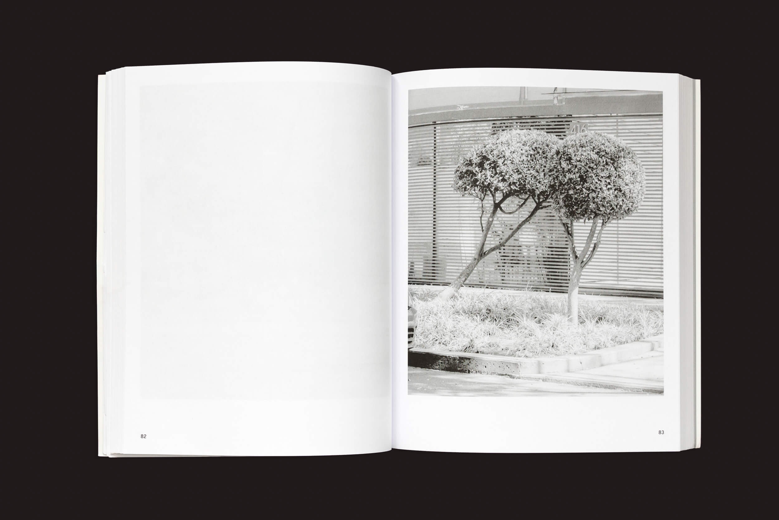 Em Rubén Montesinos: A Guide on Gardening, da Handshake, um livro aberto revela uma foto em preto e branco de uma árvore topiária bem cuidada em frente a um edifício com persianas horizontais na página direita, incorporando elegância cultural, enquanto a página esquerda permanece em branco, convidando à imaginação.