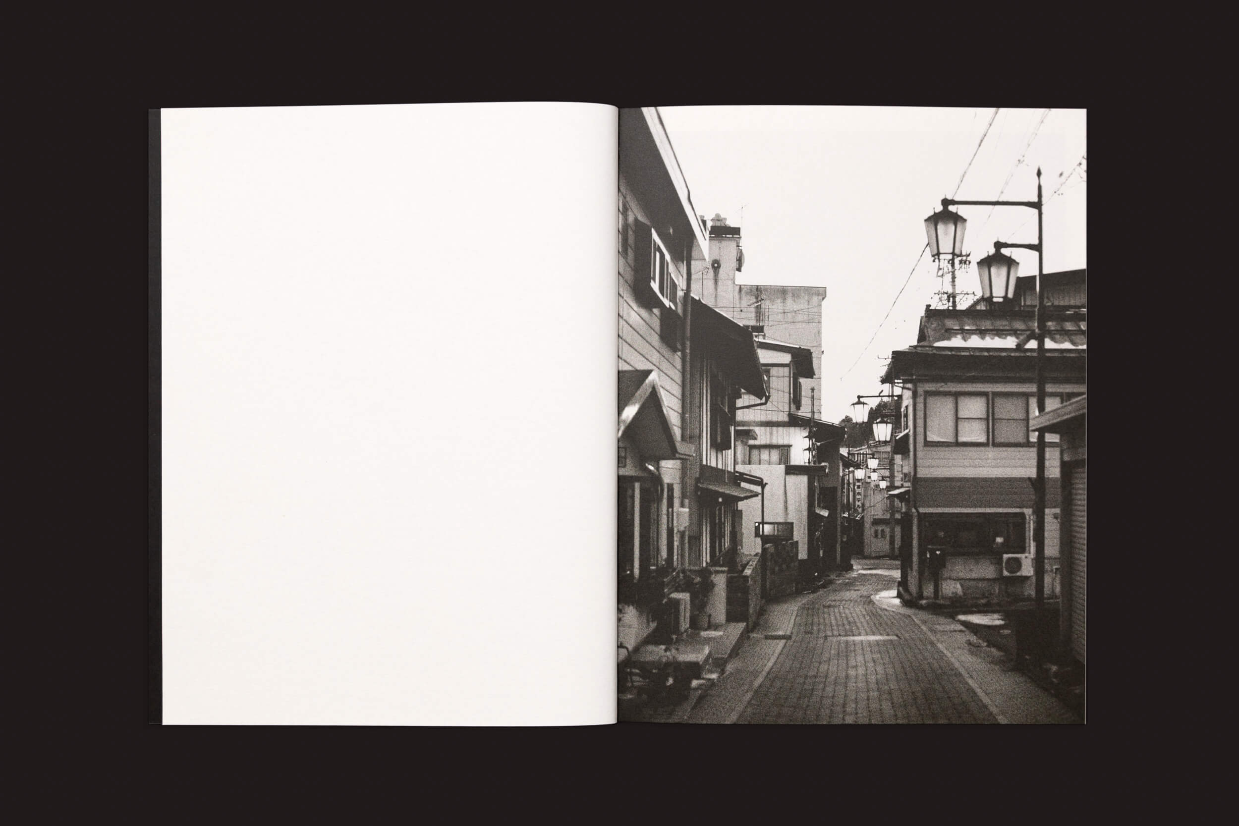 Um livro aberto exibe uma foto em preto e branco na página direita, ecoando o estilo de Layla Motta em Ainda há um longo caminho até minha casa, de Selo Turvo. Mostra uma rua estreita do Japão com edifícios e luminárias tradicionais. A página da esquerda está em branco, convidando à contemplação de uma travessia emocional.