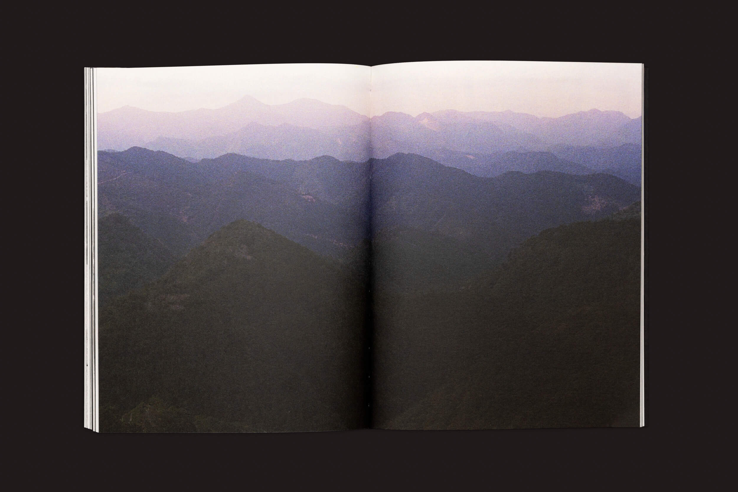 Um livro aberto mostra uma foto de uma paisagem serena em duas páginas, retratando montanhas desaparecendo sob um céu rosado. Esta cena reflete a travessia emocional de Layla Motta em sua obra Ainda há um longo caminho até minha casa de Selo Turvo, contra um fundo escuro.