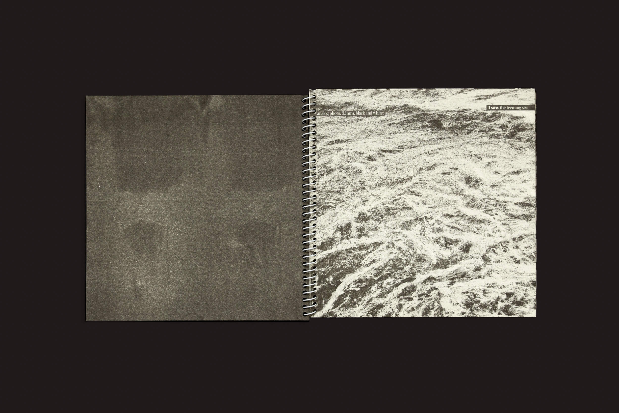 Um caderno espiral aberto repousa sobre uma superfície escura. A página da esquerda está em branco e texturizada, ideal para pensamentos criativos. A página da direita exibe Juan Manuel Lara: Aleph-2 da SED Editorial, apresentando uma textura abstrata em tons de cinza como ondas ou nuvens e um pequeno texto no topo.