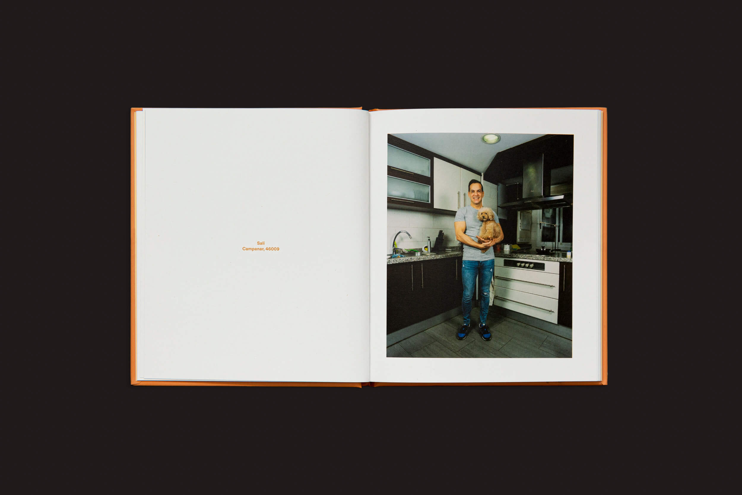 Um livro aberto repousa sobre uma superfície escura. A página da esquerda mostra um texto vermelho mínimo, enquanto na direita há uma foto de uma pessoa em uma cozinha moderna segurando o Laura Leal: Apricot Poodle da Handshake, com armários e eletrodomésticos no fundo deste ambiente aconchegante.