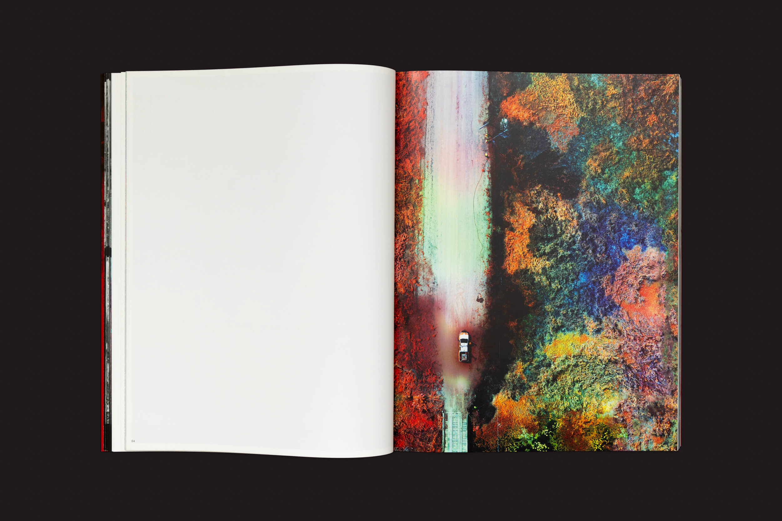 Um livro aberto com uma foto aérea brilhante e colorida de um carro em uma estrada vibrante em meio à folhagem de outono nos lembra de alterações climáticas; é Richard Mosse: Broken Spectre de Loose Joints. A página oposta está em branco, enfatizando a mensagem dura.