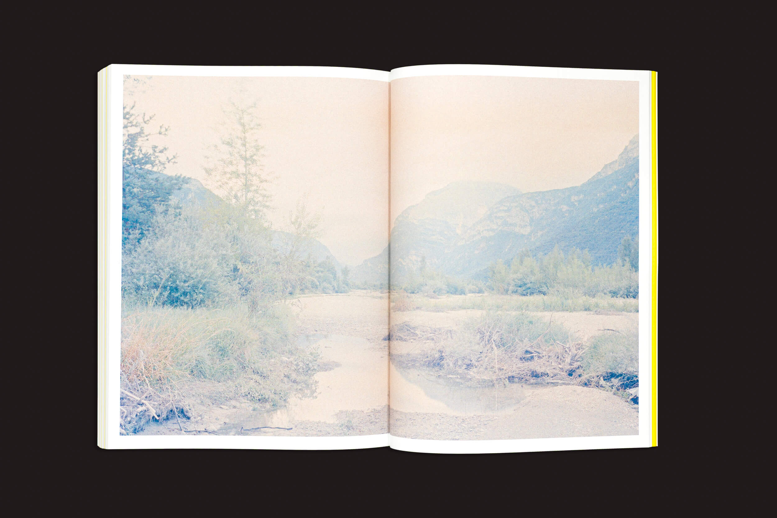 CALAMITA/À – Uma investigação sobre a catástrofe de Vajont: v/a da Fw:Books apresenta um livro aberto exibindo uma foto desbotada de uma paisagem em tons pastéis que faz uma referência sutil ao desastre de Vajont, sobre um fundo preto sólido.