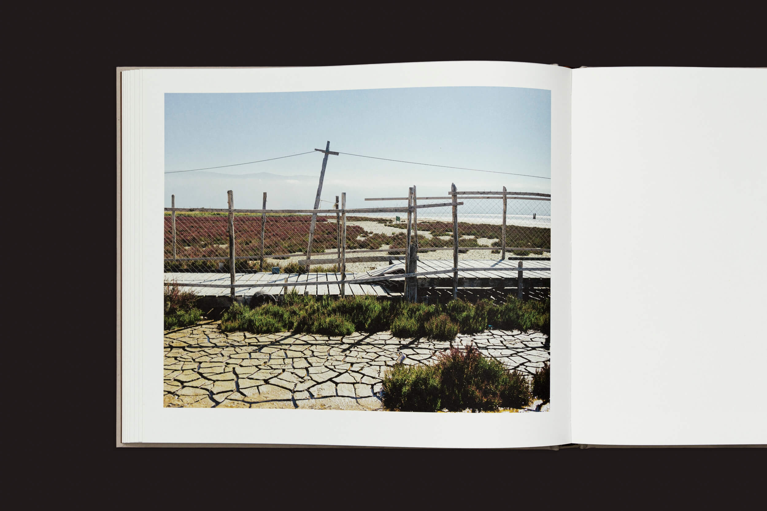 Em Caspian: A Southern Reflection de Khashayar Javanmardi da Loose Joints, um livro aberto exibe uma foto de uma paisagem seca e rachada com vegetação esparsa. Uma cerca de arame e postes de madeira ficam sob um céu azul claro, contrastando fortemente com o fundo escuro, sugerindo o impacto da mudança climática.