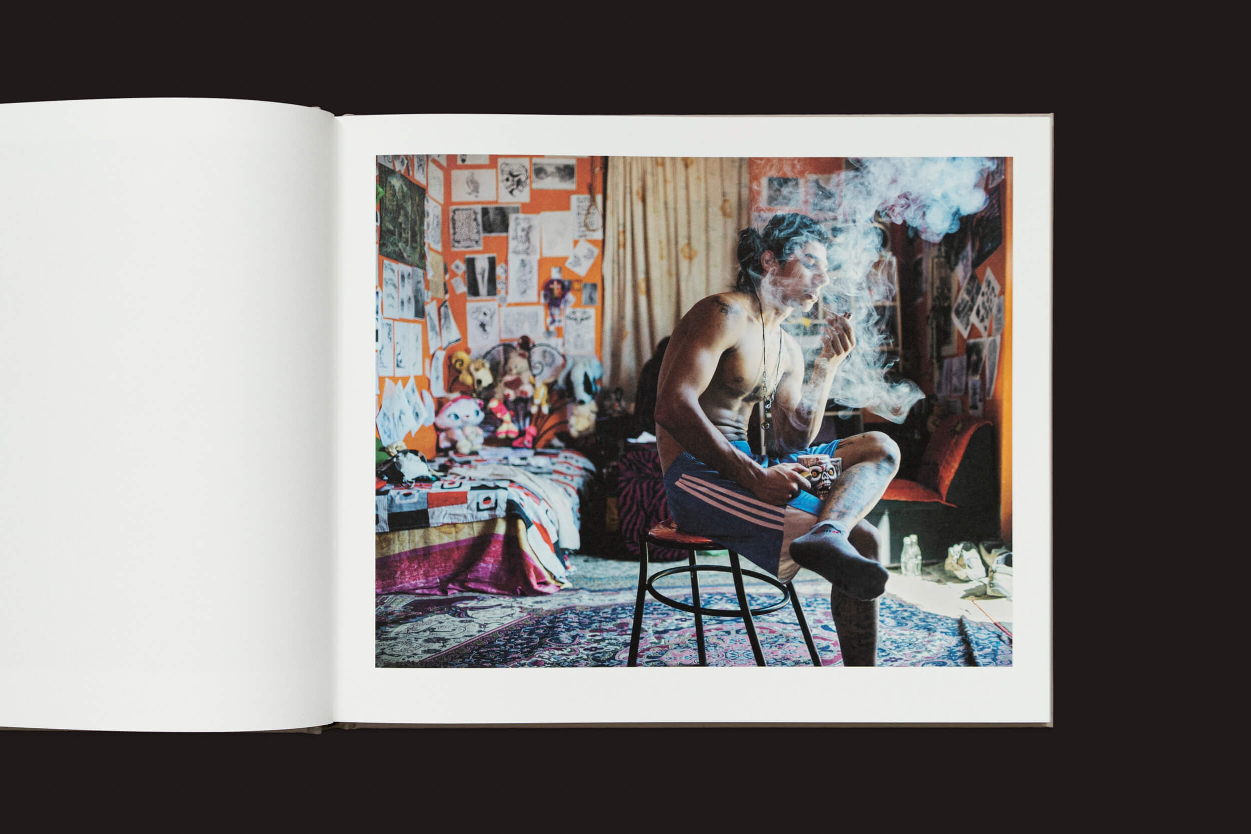 Um homem relaxa sem camisa em um banco em uma sala desorganizada de Caspian: A Southern Reflection de Khashayar Javanmardi (Loose Joints), exalando fumaça. Arte, pôsteres, bichos de pelúcia e um tapete estampado criam uma atmosfera aconchegante, apesar da poluição ambiental.