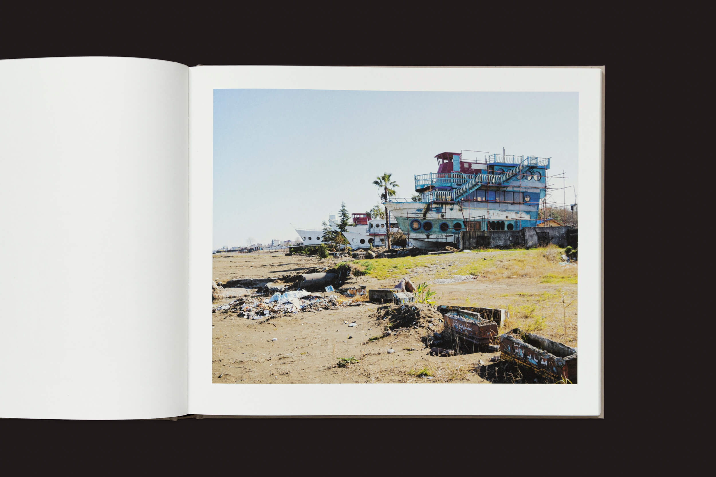 Em Khashayar Javanmardi: Caspian: A Southern Reflection de Loose Joints, um livro aberto apresenta uma foto de um edifício dilapidado, semelhante a um navio, com janelas de vigia e tinta descascada. Terra árida e coberta de detritos em primeiro plano, sob um céu limpo, reflete a poluição ambiental.