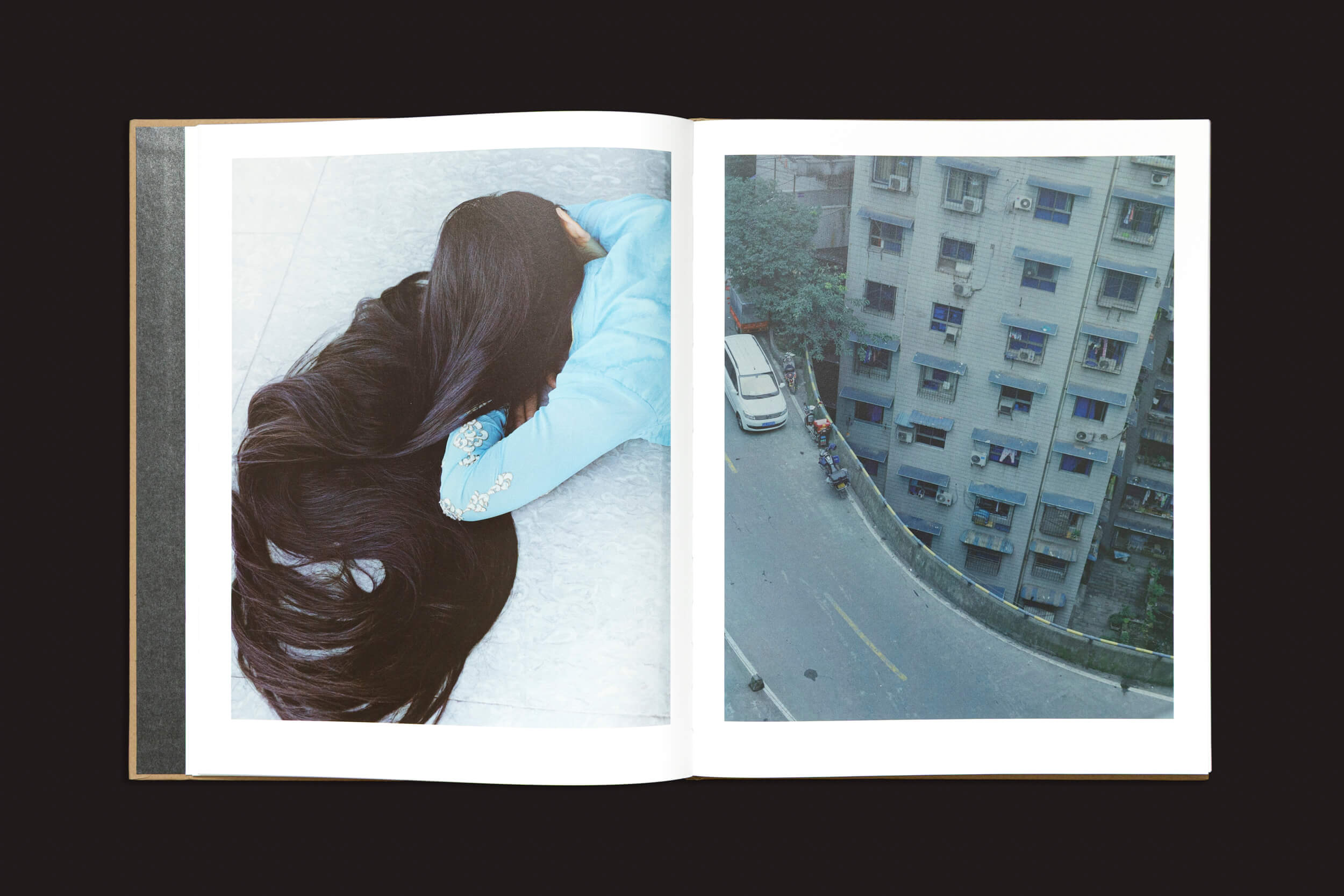 Um livro aberto exibe duas páginas: a da esquerda apresenta uma pessoa com longos cabelos negros e roupas azuis, simbolizando a identidade fragmentada; a da direita mostra uma vista aérea de uma rua da cidade que lembra o foco da diáspora de segunda geração de Teresa Eng, de "Teresa Eng: China Dream", de Skinnerboox.