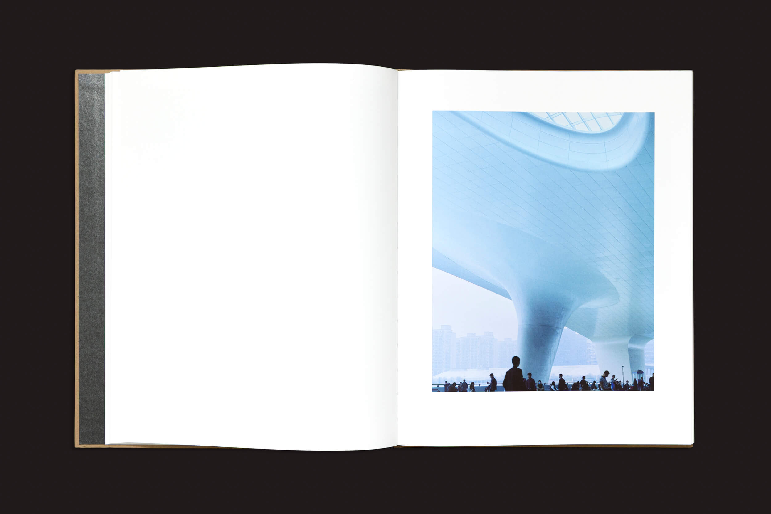 Um livro aberto, "Teresa Eng: China Dream", de Skinnerboox, mostra uma foto de um interior arquitetônico com um teto curvo futurista. Figuras em silhueta acrescentam escala, alinhando-se ao tema de identidade fragmentada de Eng, enquanto a página em branco convida à reflexão.