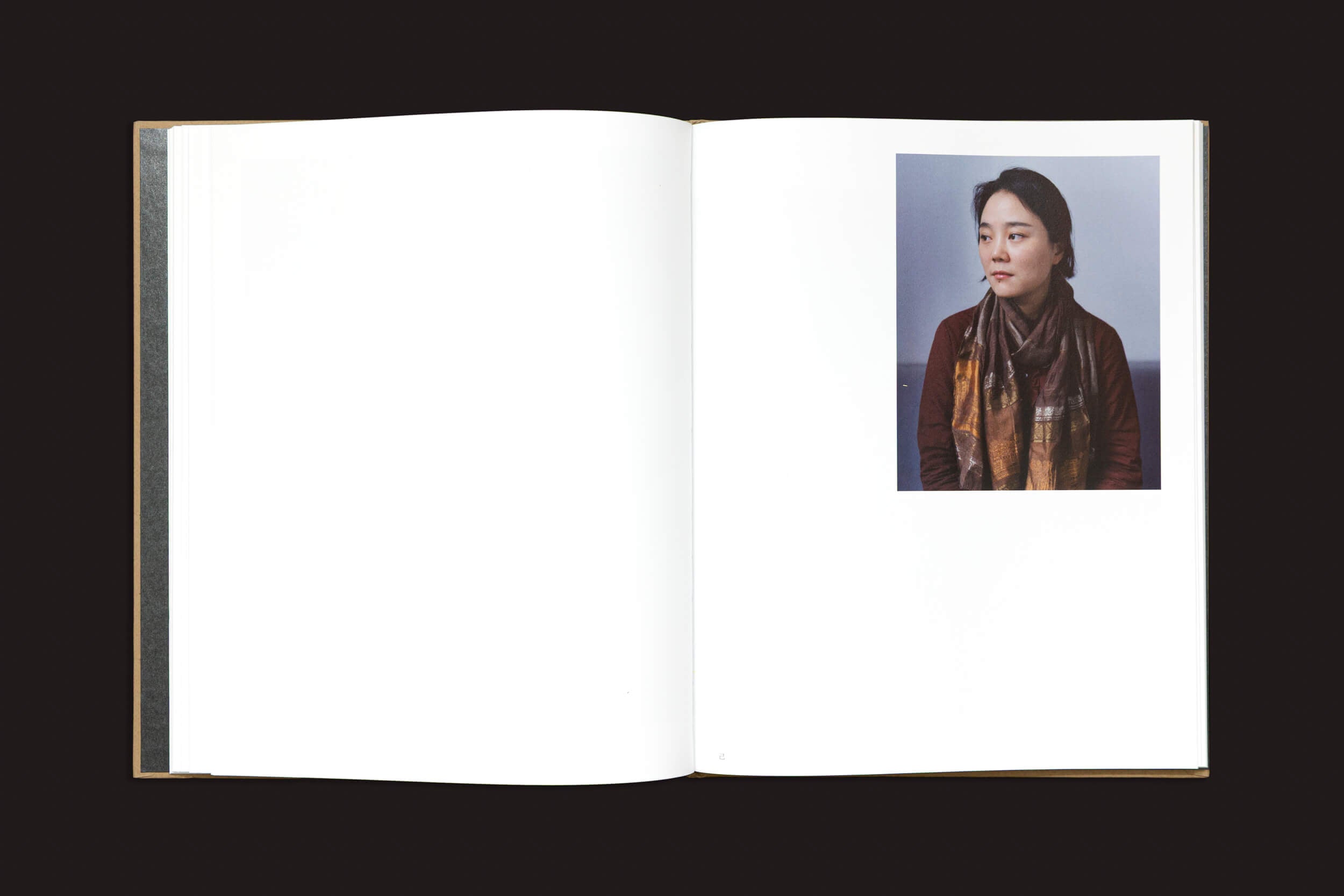 Um livro aberto com páginas em branco apresenta um retrato à direita: uma pessoa de cabelos escuros, com jaqueta e cachecol marrons, olhando para a direita contra um fundo simples. Um sussurro visual de identidade fragmentada em "China Dream", de Teresa Eng, por Skinnerboox.