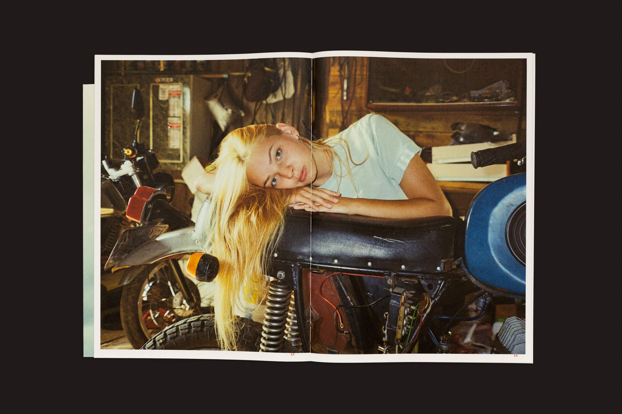 Uma mulher com longos cabelos loiros se inclina sobre um assento de motocicleta, vestindo uma camisa azul-clara. A garagem mal iluminada, lembrando uma cena de Toma Gerzha: Control Refresh, da The Eriskay Connection, está cheia de ferramentas e peças de reposição espalhadas ao fundo.