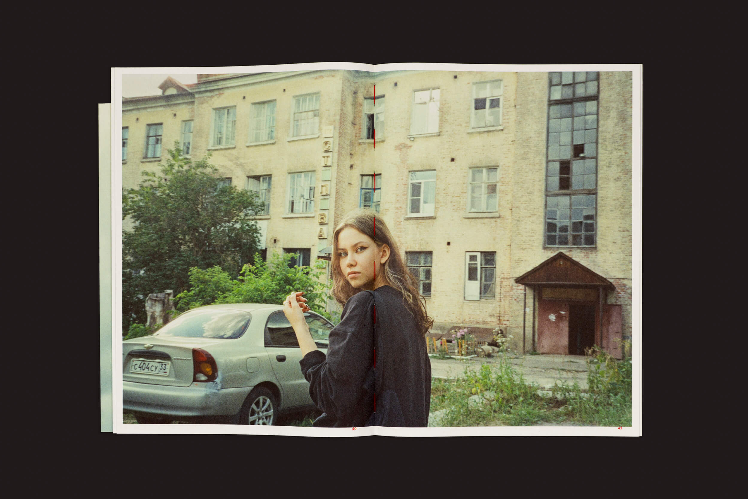 Um indivíduo de cabelos longos com uma jaqueta escura está diante de um prédio grande e envelhecido, segurando um objeto. Perto dali, um carro prateado está estacionado. A cena evoca Toma Gerzha: Control Refresh de The Eriskay Connection, capturando a essência da idade e da negligência.