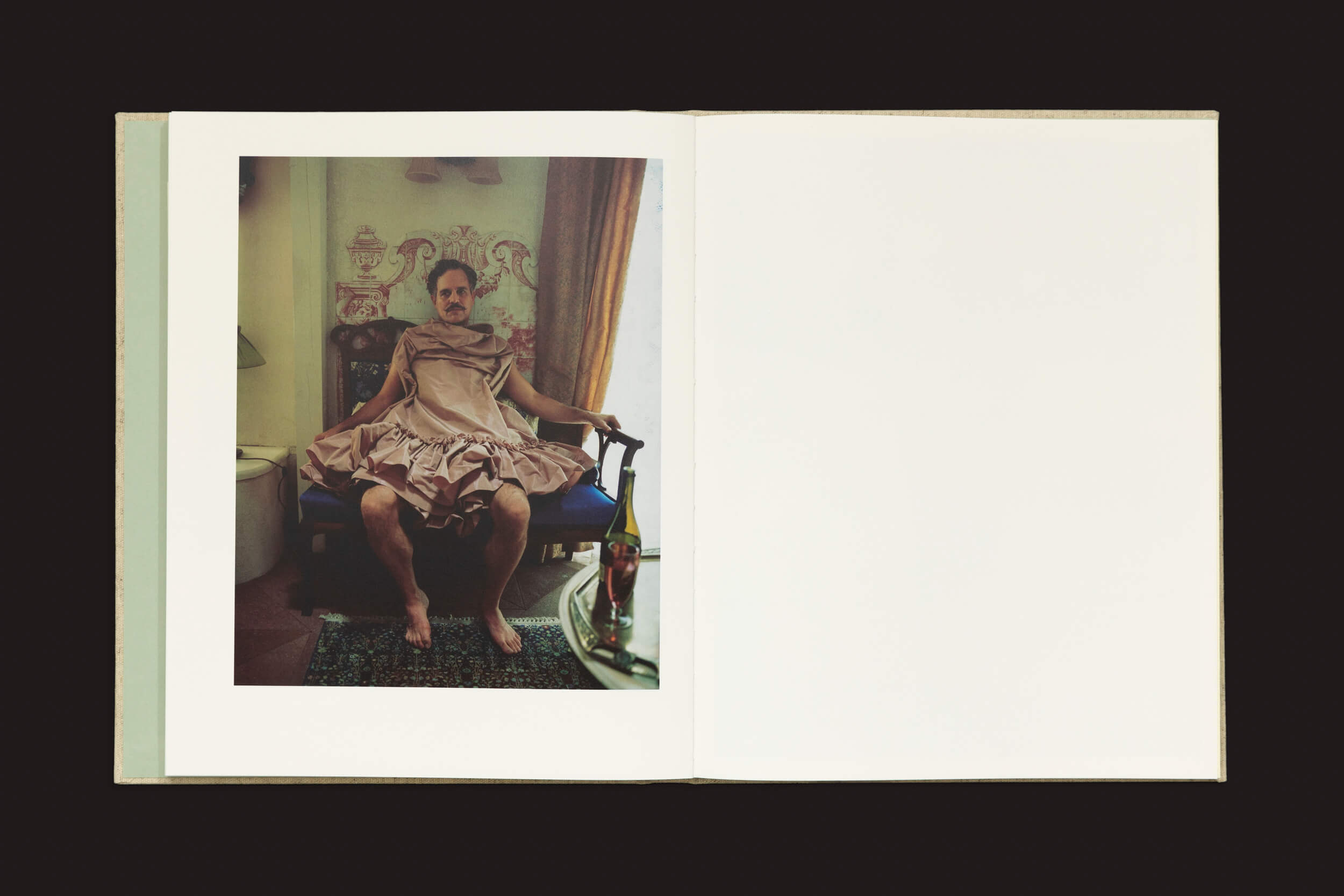 Um livro aberto mostra uma foto do século XIX à esquerda, retratando uma pessoa descalça em um vestido de babados sentada em uma cadeira. A página da direita está em branco, com um ambiente de Yorgos Lanthimos: Querido Deus, o Partenon ainda está quebrado pelo Vazio — papel de parede estampado e vislumbres de vinho completam a cena.