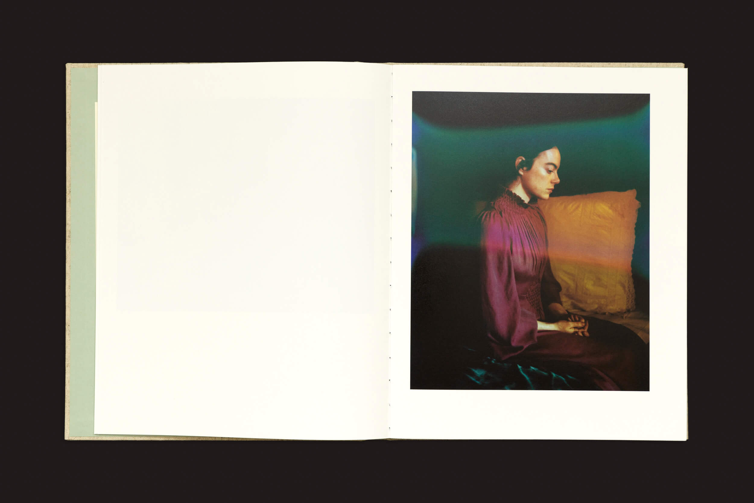 Um livro aberto intitulado Yorgos Lanthimos: Dear God, the Parthenon is still broken de Void revela uma foto na página da direita. Ela mostra uma pessoa em um vestido rosa sentada de perfil com as mãos entrelaçadas contra um cenário sombrio iluminado por vibrantes tons de verde e amarelo. A página da esquerda está quase toda em branco.