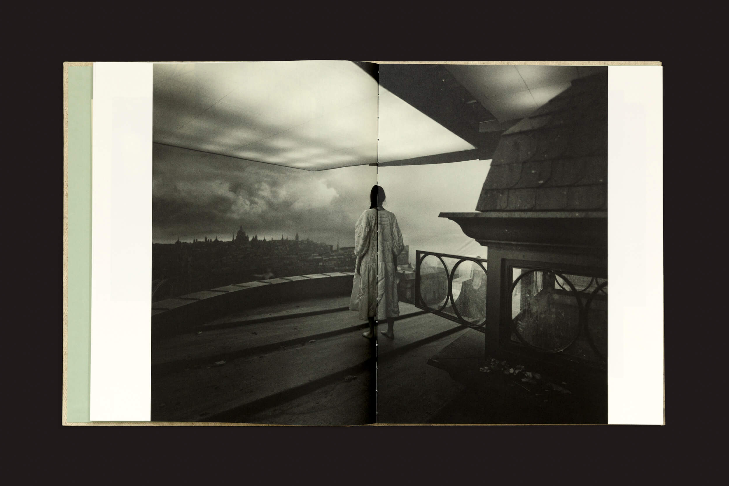 Uma pessoa com um casaco longo está em um telhado, com vista para uma paisagem urbana que lembra Yorgos Lanthimos: Dear God, the Parthenon is still broken de Void. A cena em preto e branco, com nuvens dramáticas, evoca a solidão encontrada nas fotografias fotográficas do século XIX.