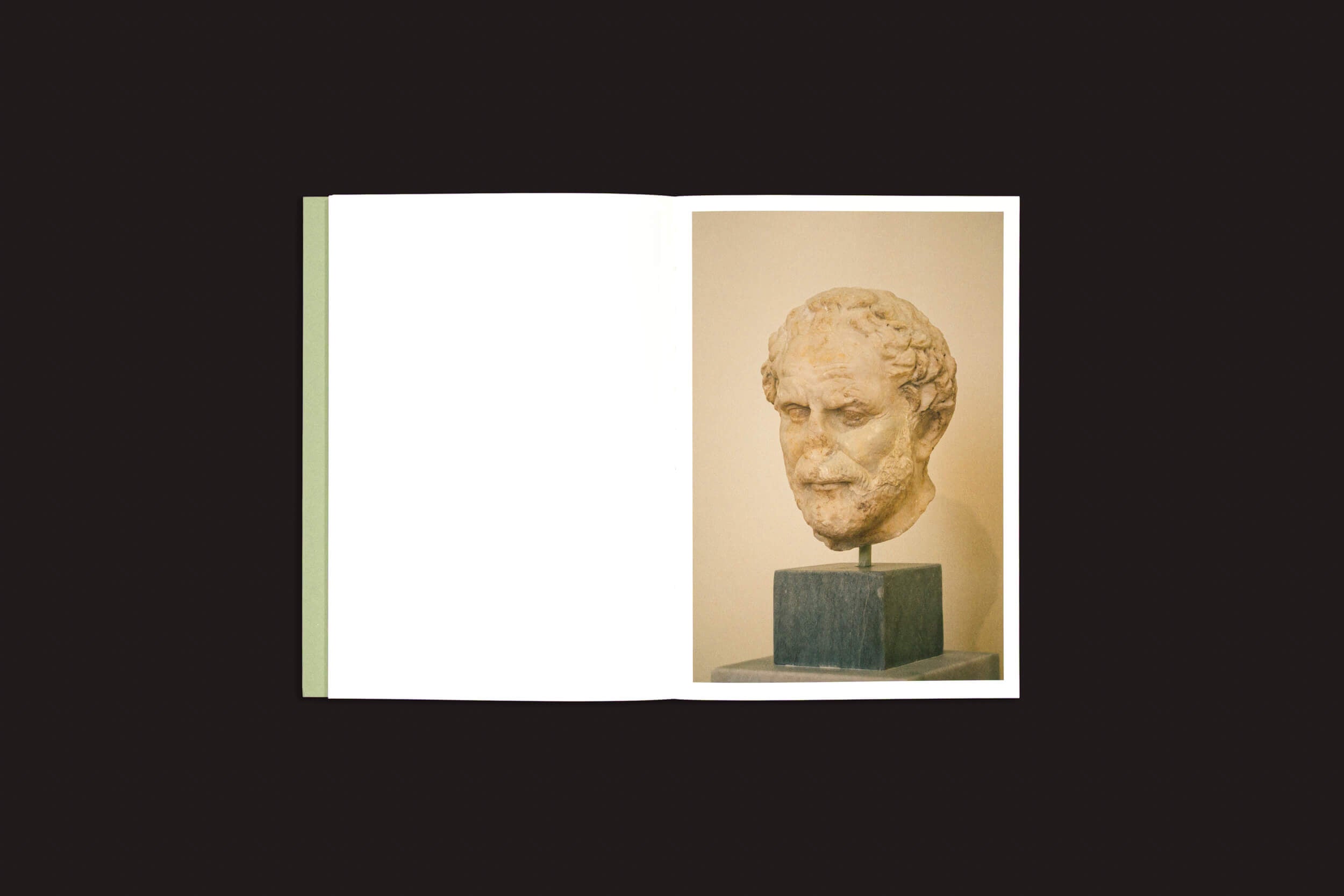 Contra um fundo escuro, um livro aberto intitulado "Simon Bray: Caro Kairos", de Skinnerboox, mostra a foto de um antigo busto de pedra com características faciais detalhadas na página direita. A página esquerda permanece em branco, como se aguardasse os pensamentos do Caro Kairos.