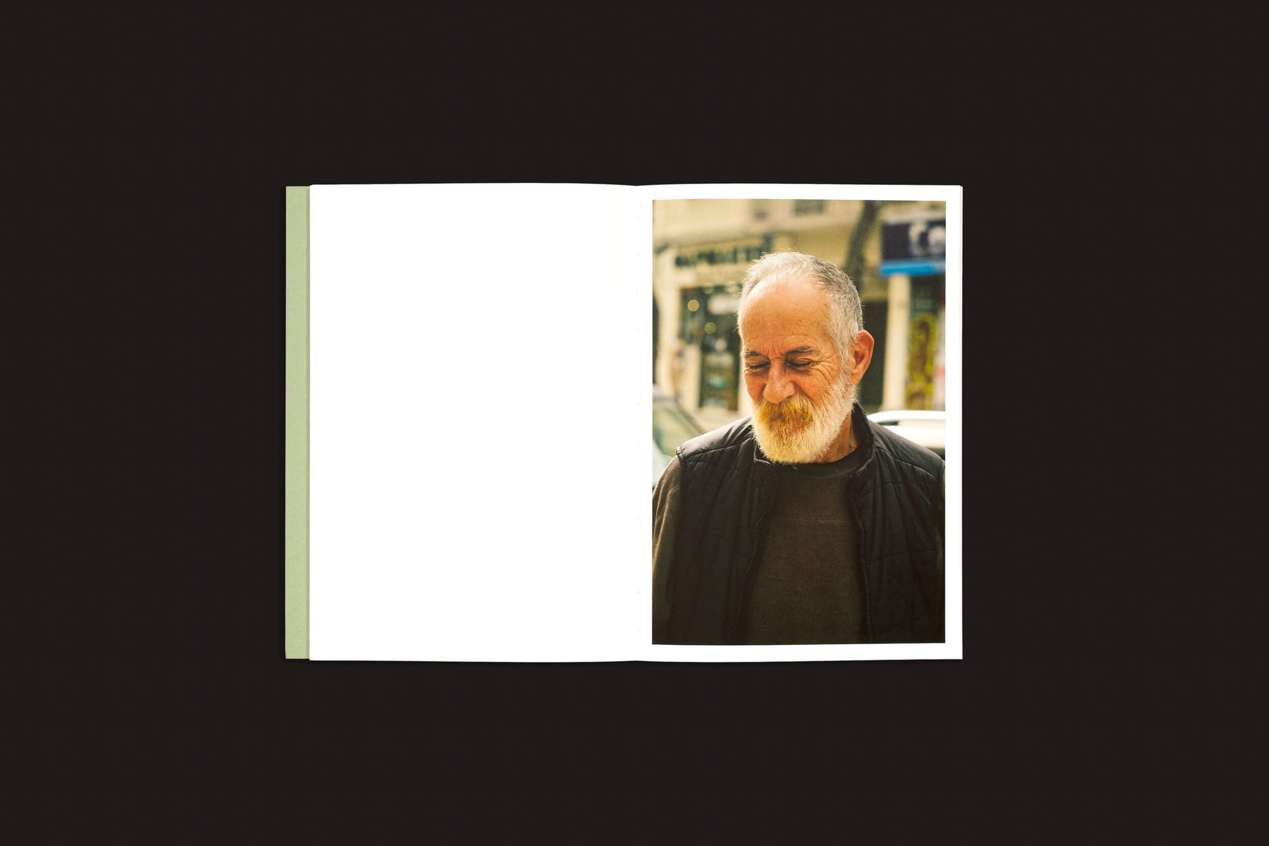 O fundo preto emoldura "Simon Bray: Dear Kairos", de Skinnerboox. A página da esquerda está em branco, simbolizando o tempo não escrito; a da direita exibe um homem barbudo de paletó escuro olhando para uma rua de Atenas, capturando um momento com a graça do tempo grego.