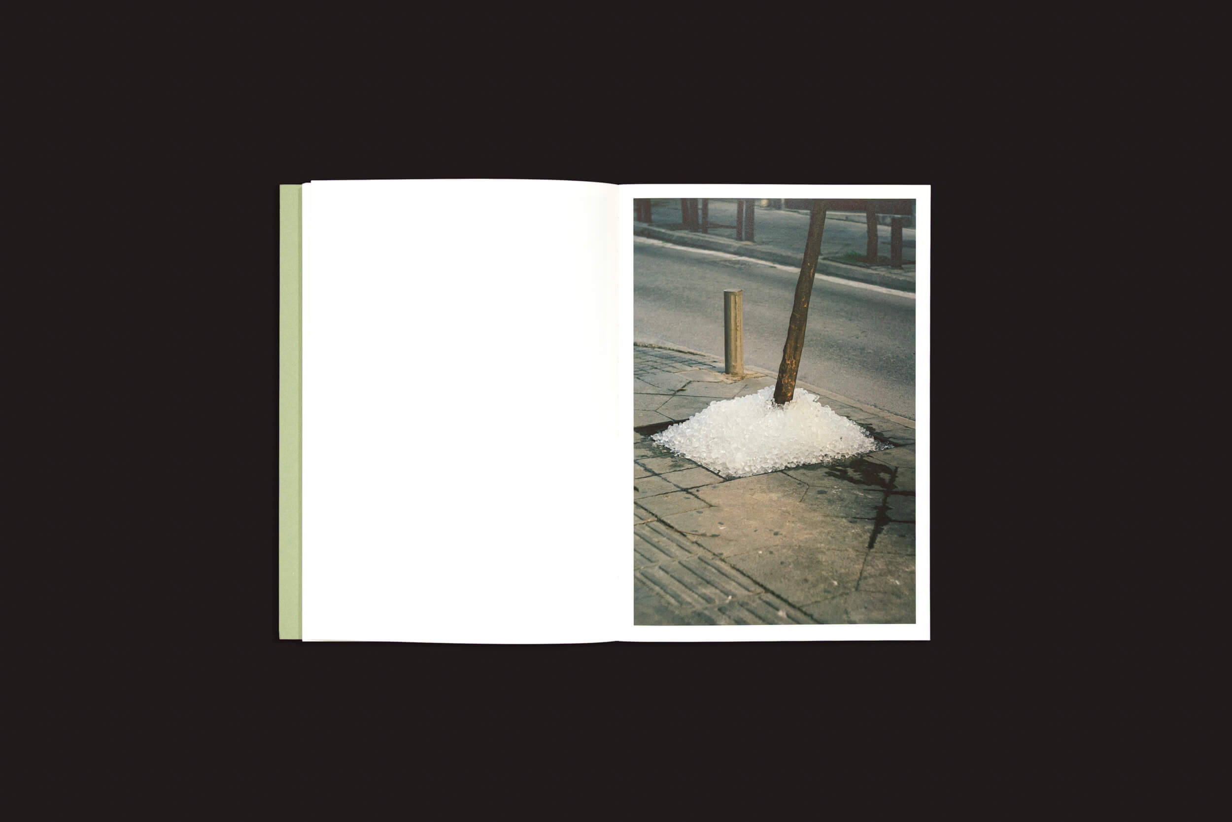 Um exemplar aberto de "Simon Bray: Dear Kairos", de Skinnerboox, repousa sobre uma superfície escura. A página da direita revela uma cena de rua em Atenas, enquanto a da esquerda permanece em branco, ecoando o fascínio atemporal do tempo grego.