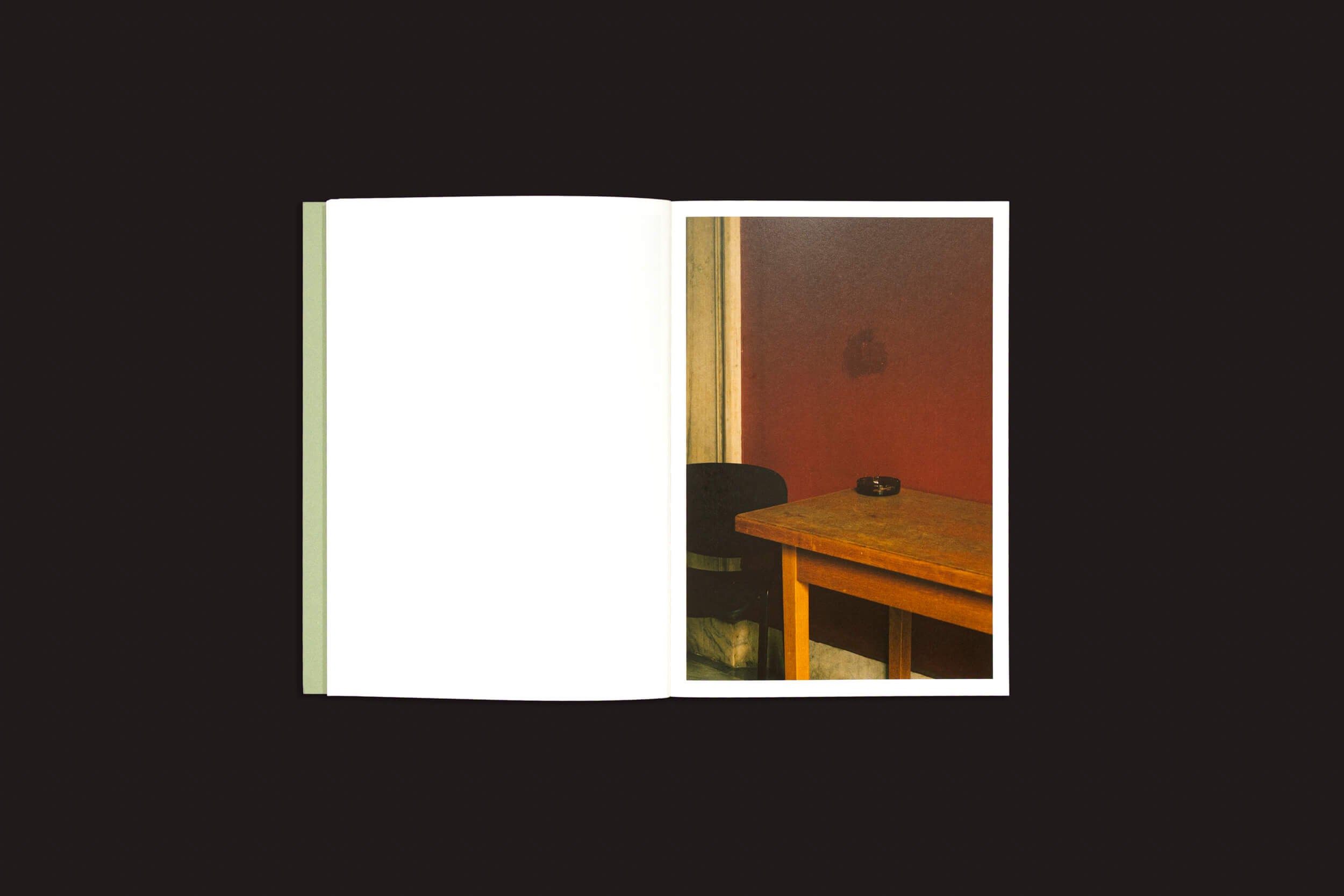 Um livro aberto revela a foto de uma mesa de madeira encostada em uma parede vermelha. Sobre a mesa, um cinzeiro, com parte de uma cadeira preta visível. A página da esquerda permanece em branco — uma sutil referência a "Simon Bray: Dear Kairos", de Skinnerboox — sobre um fundo preto, capturando o ritmo serendipitário da vida.