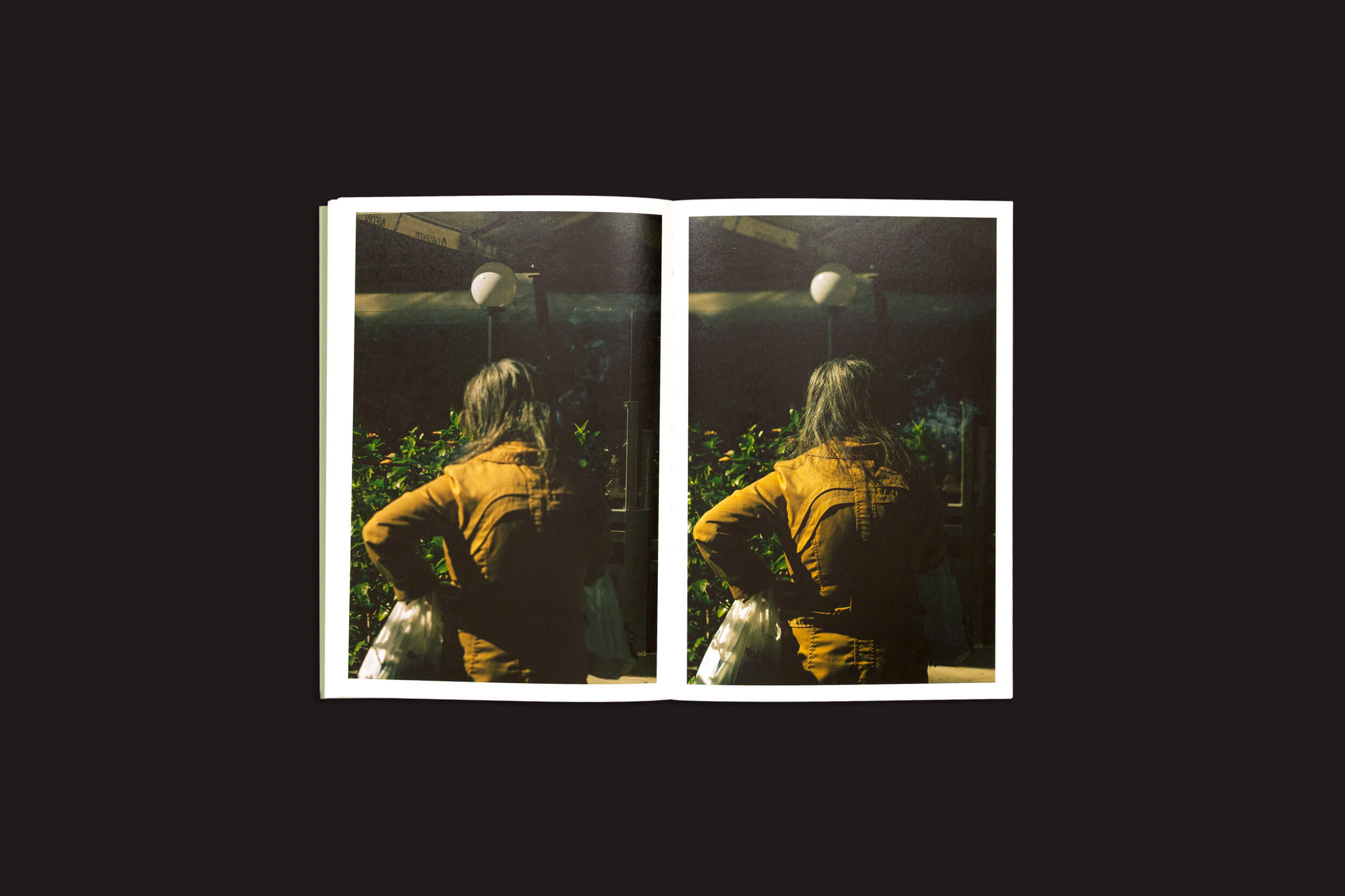 Um exemplar de Simon Bray: Dear Kairos, de Skinnerboox, mostra uma pessoa de cabelos longos, de jaqueta marrom, com as mãos na cintura, contemplando a vegetação de Atenas e o cenário mal iluminado. Uma luz circular realça a atmosfera de tempo grego.