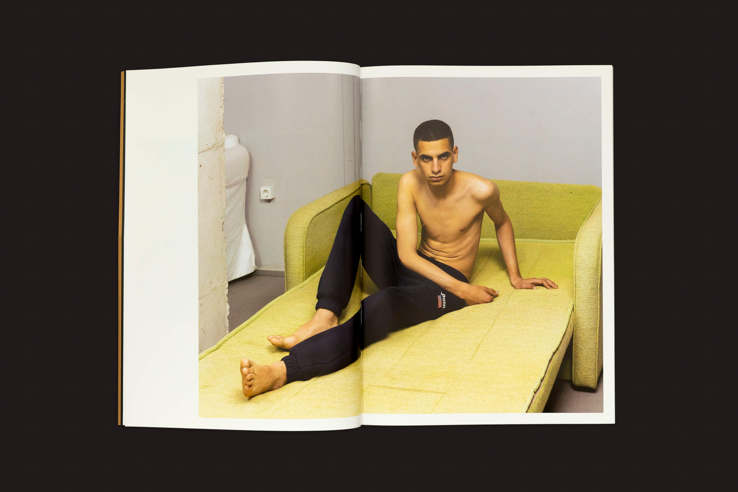 Em uma revista aberta, uma imagem surreal de Felipe Romero Beltrán: Dialect de Loose Joints mostra um homem sem camisa em um sofá verde. Suas pernas se estendem por duas páginas, criando uma ilusão alongada contra um fundo escuro, espelhando sutilmente o contraste imponente que lembra nuances legais.