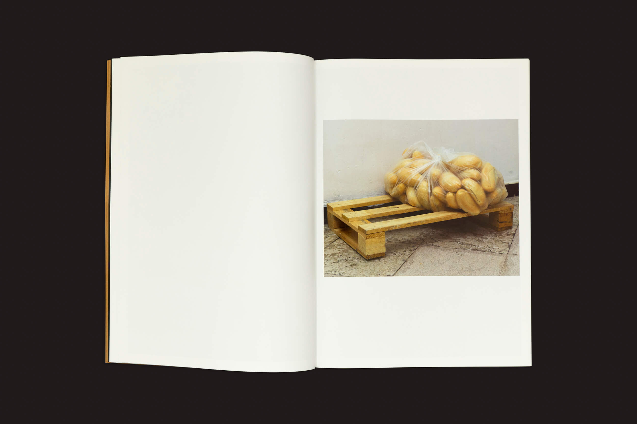 O livro aberto revela uma fotografia de um palete de madeira com um saco transparente cheio de pãezinhos redondos, capturando simplicidade e calor. Contra o fundo escuro, Dialect de Felipe Romero Beltrán, de Loose Joints, sussurra histórias não contadas de estatuto legal e jornadas de imigrantes marroquinos.