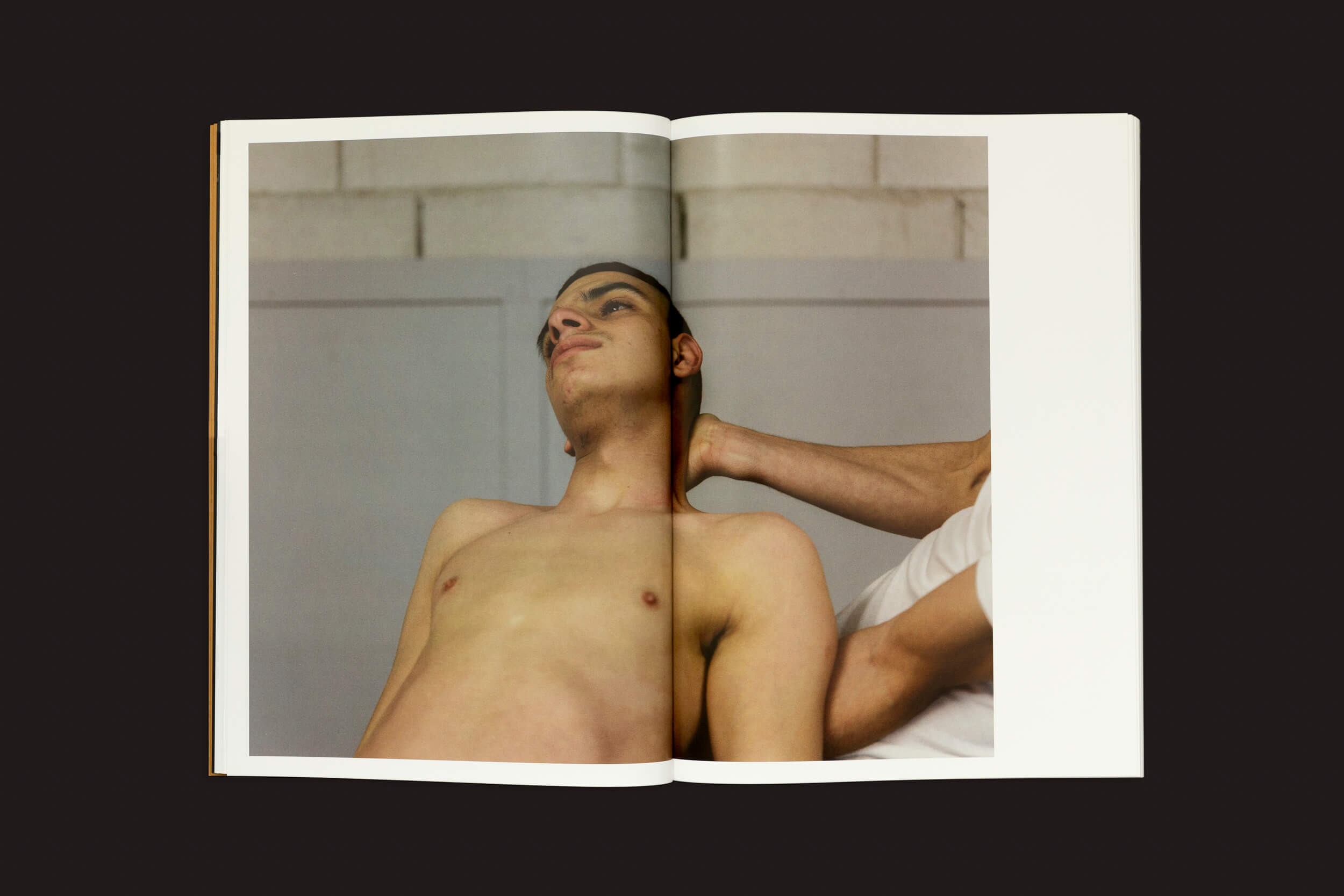 Em Dialect, de Felipe Romero Beltrán, publicado pela Loose Joints, um livro aberto revela uma foto comovente de uma pessoa sem camisa reclinada, apoiada pela mão de outra pessoa em seu pescoço, em um cenário neutro que evoca temas de resiliência e vulnerabilidade frequentemente enfrentados por imigrantes marroquinos.