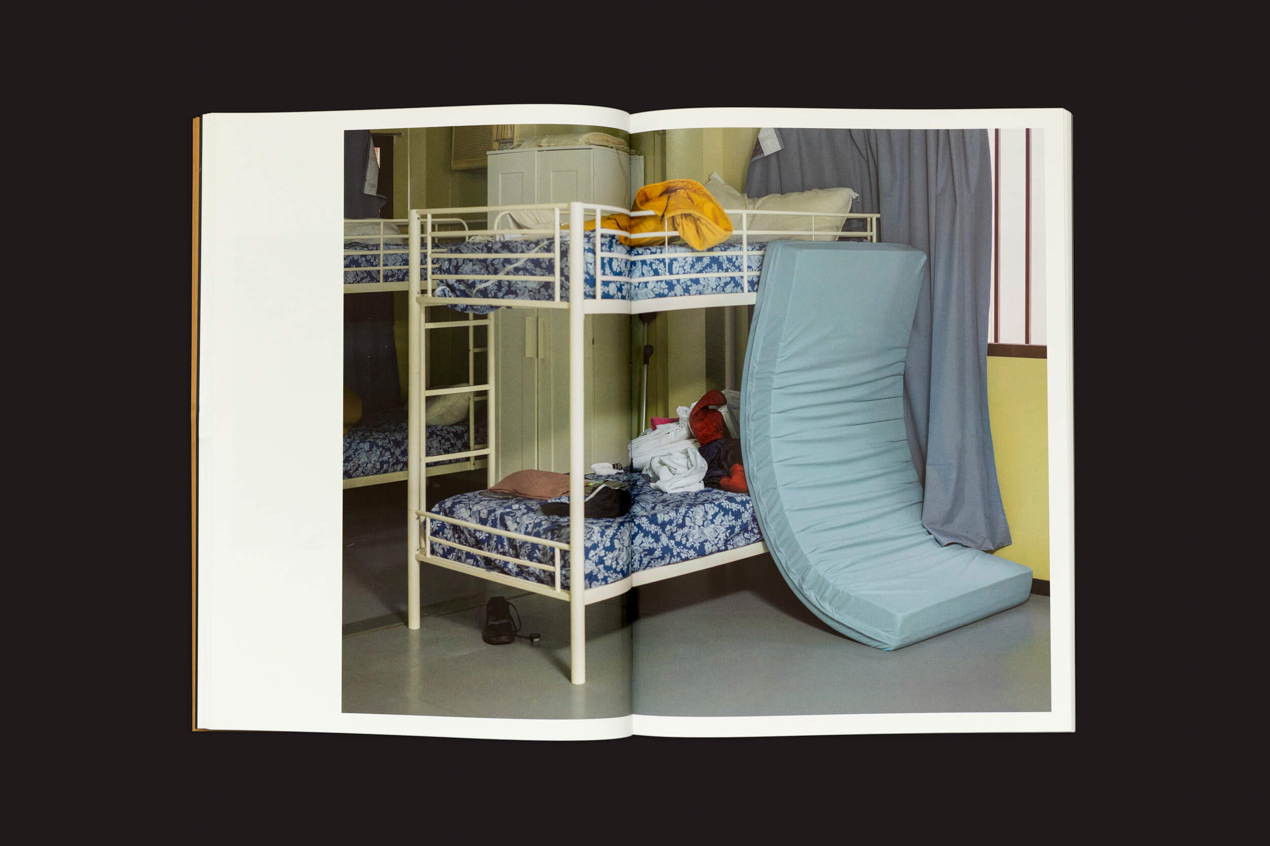 Um livro aberto exibe uma imagem de Felipe Romero Beltrán: Dialect de Loose Joints, apresentando um beliche com roupa de cama estampada em azul. Roupas cobrem o beliche de cima, a parte de baixo segura itens e um colchão dobrado se inclina próximo, sugerindo movimento ou transição. Um armário e cortinas completam a cena.