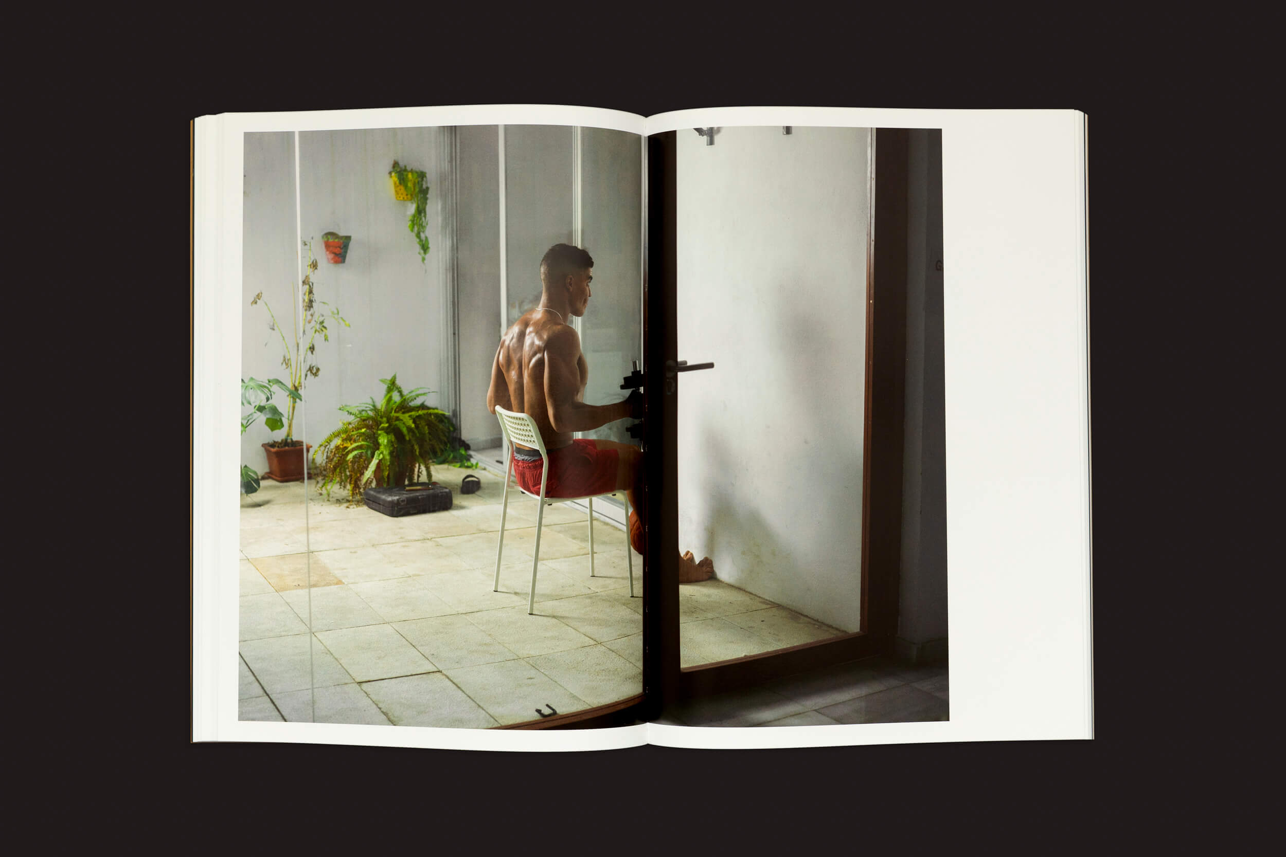 Dentro do livro aberto Felipe Romero Beltrán: Dialect de Loose Joints, uma fotografia serena captura uma pessoa sem camisa, de shorts vermelho, sentada em uma cadeira dentro de casa, cercada por plantas. Essa cena calma contrasta com histórias de estatuto legal severo enfrentadas por imigrantes marroquinos.