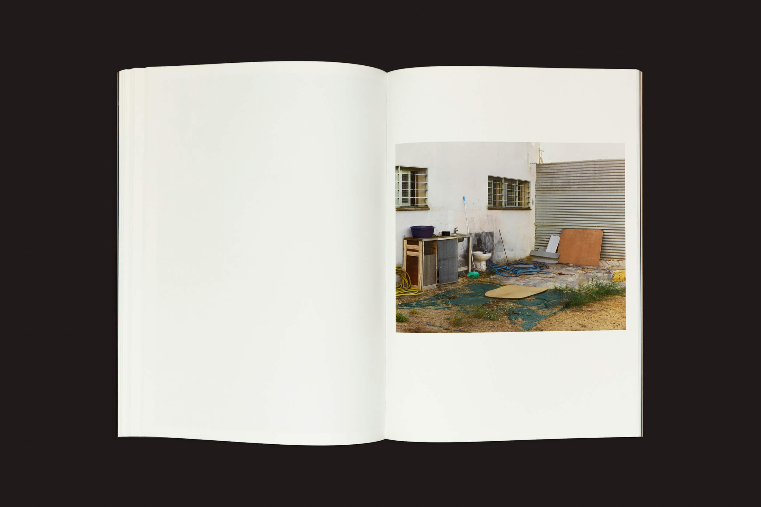 Em uma superfície escura, um livro aberto exibe uma foto de uma cena ao ar livre — paredes brancas com janelas gradeadas, tábuas espalhadas e mangueiras em meio à grama e à sujeira. Ela sugere imigrantes marroquinos em contextos de violência estatal. Esta imagem é de Felipe Romero Beltrán: Dialect de Loose Joints. As páginas estão quase todas em branco.