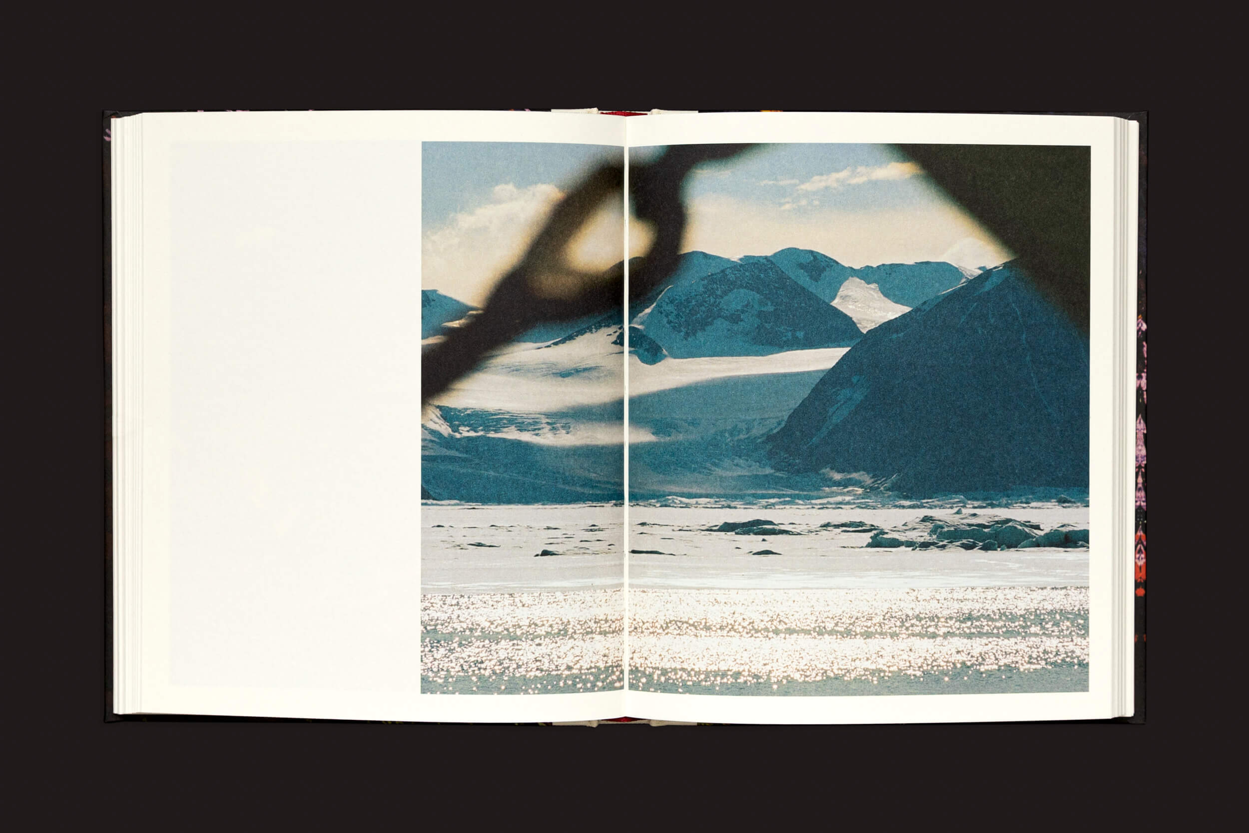 O livro Pia Riverola: Días de Loose Joints está aberto, exibindo imagens imaginativas de paisagens geladas e montanhas cobertas de neve. Um galho ou forma escura obscurece parte da visão ao longo da dobra, enquanto um fundo escuro sólido contrasta para aprimorar a experiência sensorial.