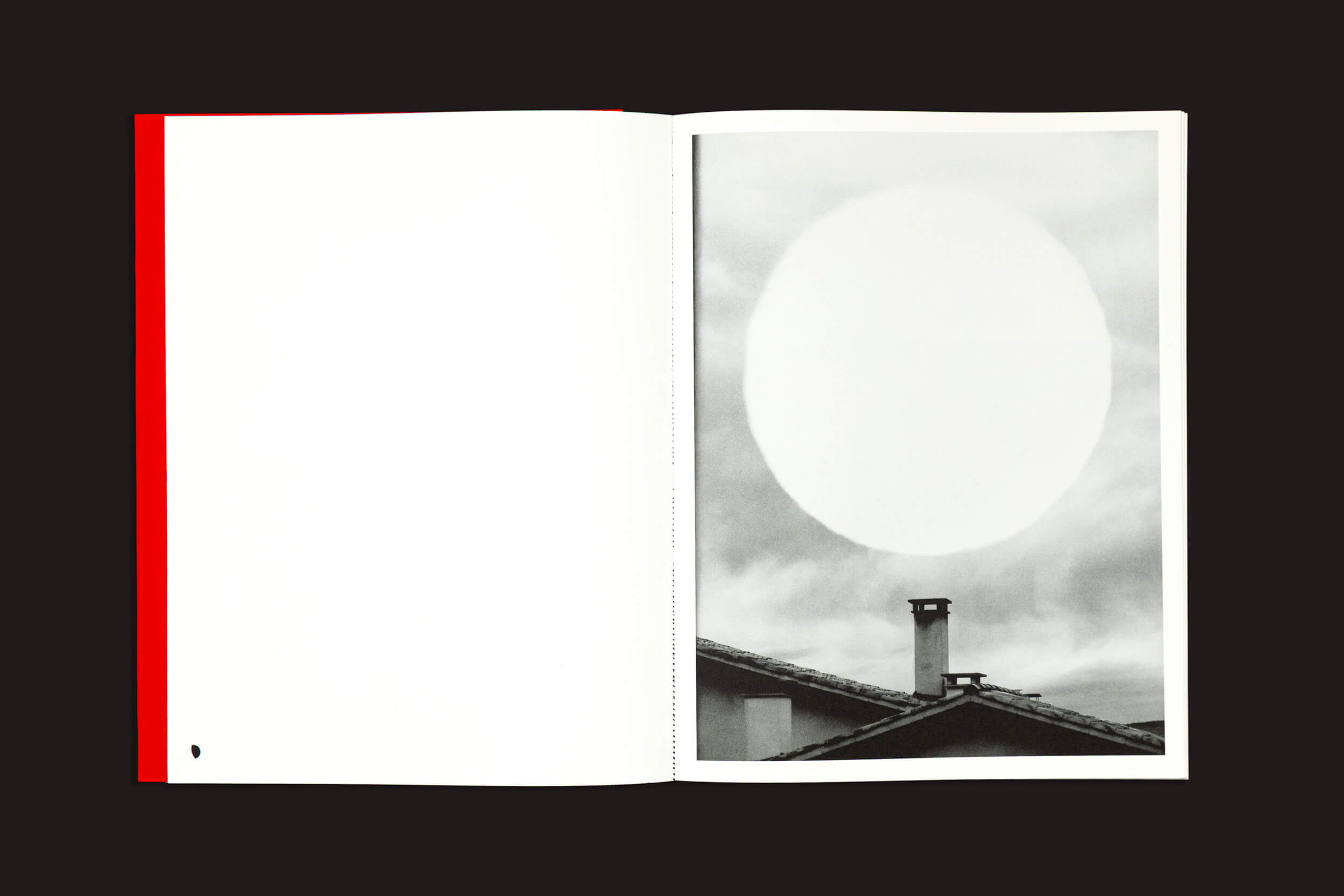 Em exposição, um livro aberto intitulado "Dream Moons", de Yurian Quintanas Nobel, da marca Void. Na página da direita, há uma fotografia em preto e branco mostrando uma grande figura circular sobre um telhado sob um céu nublado. A página da esquerda permanece em branco, com uma pequena marca preta.