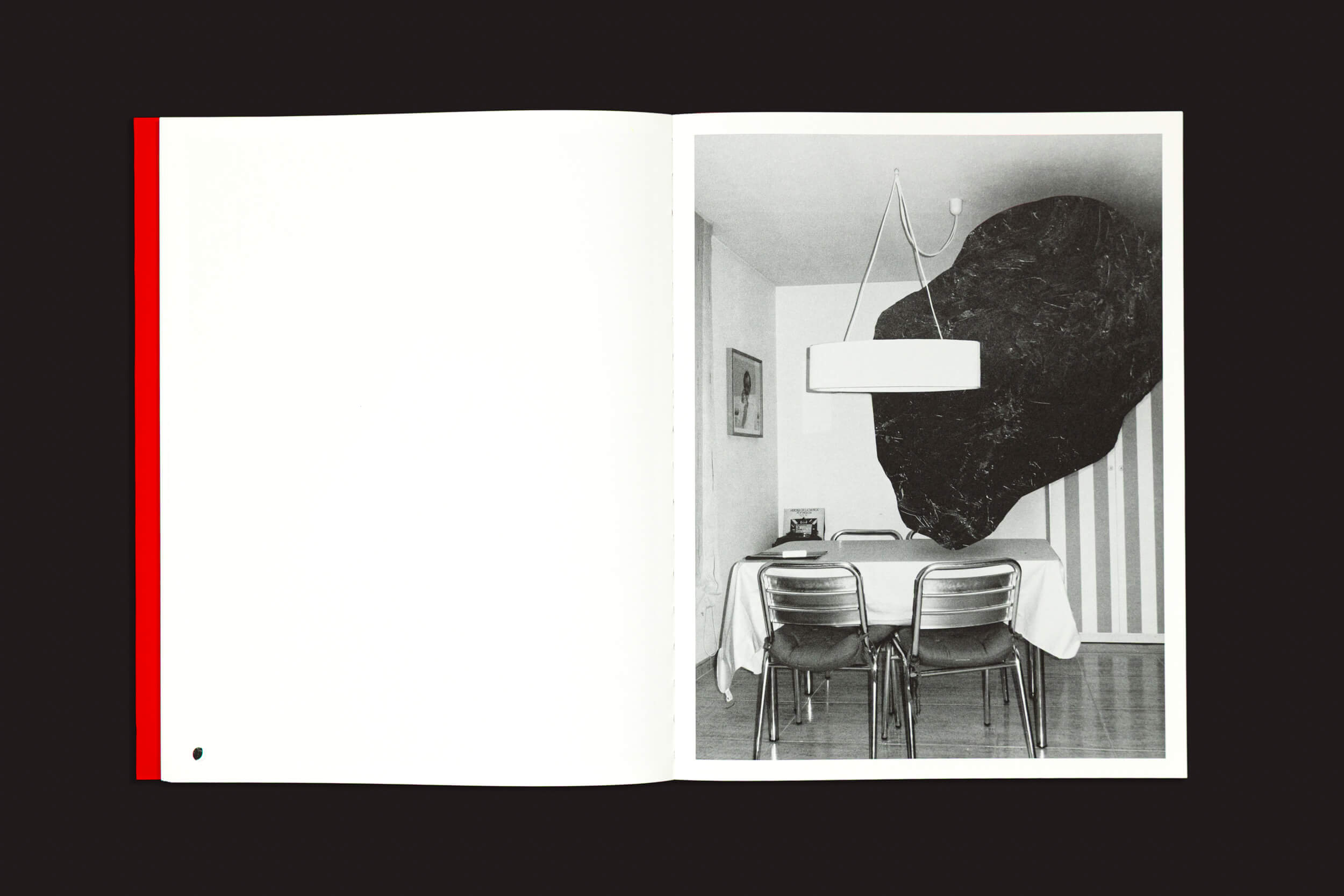 Livro aberto com uma página em branco à esquerda e uma foto em preto e branco que lembra "Dream Moons", de Yurian Quintanas Nobel, à direita, Void mostra uma mesa de jantar com três cadeiras, um objeto de parede escuro e abstrato e um pendente que ilumina essa cena surreal.