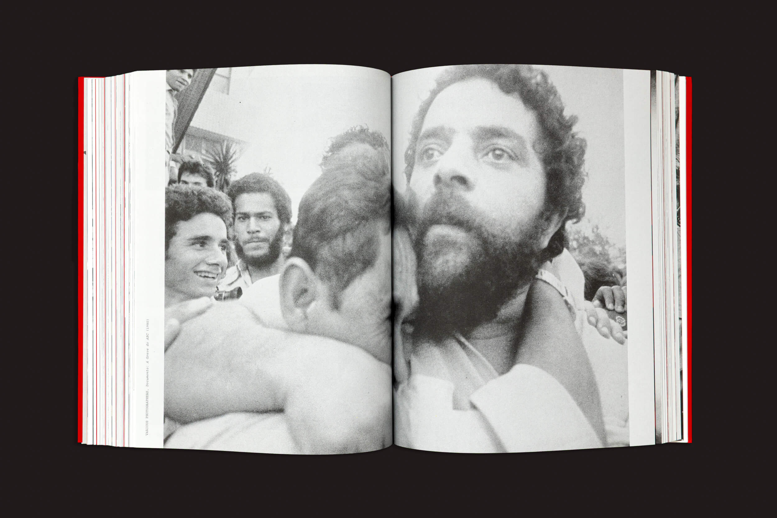 Um exemplar de "FLASHPOINT! Protest Photography in Print, 1950–Present", de Russet Lederman e Olga Yatskevich, publicado pela 10x10 Photobooks, está aberto em uma página que mostra uma fotografia em preto e branco de um homem de cabelo cacheado e barba abraçando outro, tendo como pano de fundo figuras sorridentes que personificam a resistência.
