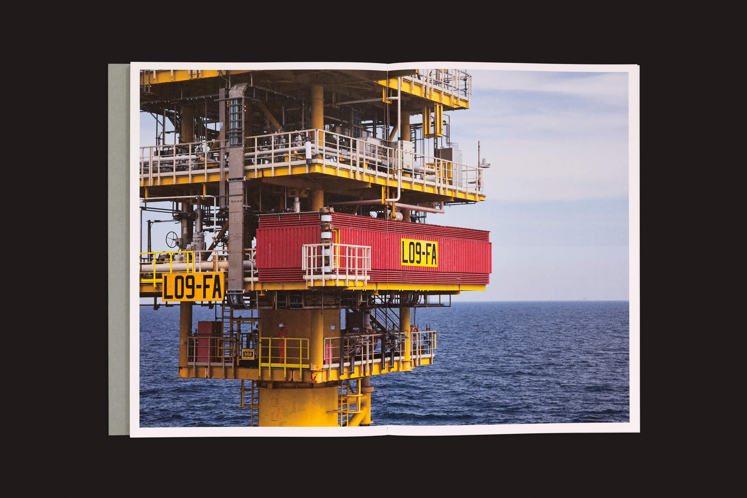 Em Tanja Engelberts: Forgotten Seas da The Eriskay Connection, um livro aberto revela uma imagem cativante de uma plataforma de petróleo offshore contra um sereno oceano azul. A estrutura amarela apresenta uma seção vermelha em negrito rotulada L09-FA, simbolizando o espírito da indústria de petróleo e gás sob céus parcialmente nublados.