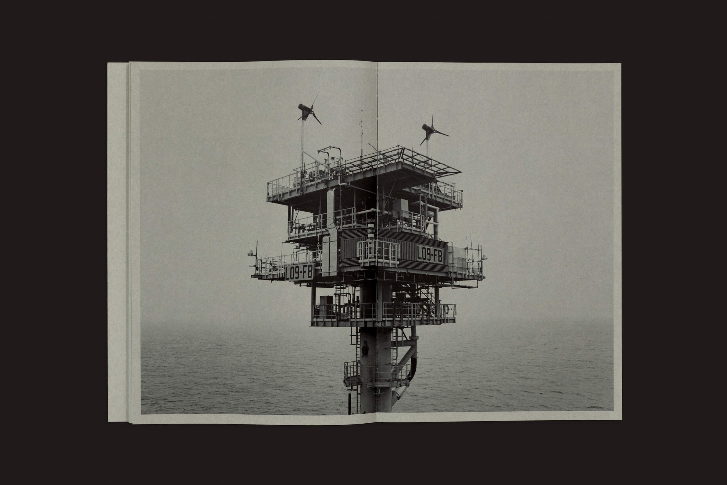 Uma imagem em preto e branco de uma plataforma offshore solitária, representando a indústria de petróleo e gás, impressa em Tanja Engelberts: Forgotten Seas por The Eriskay Connection. A plataforma apresenta vários níveis com grades, duas pequenas turbinas eólicas no topo e um mar infinito sob um céu branco.