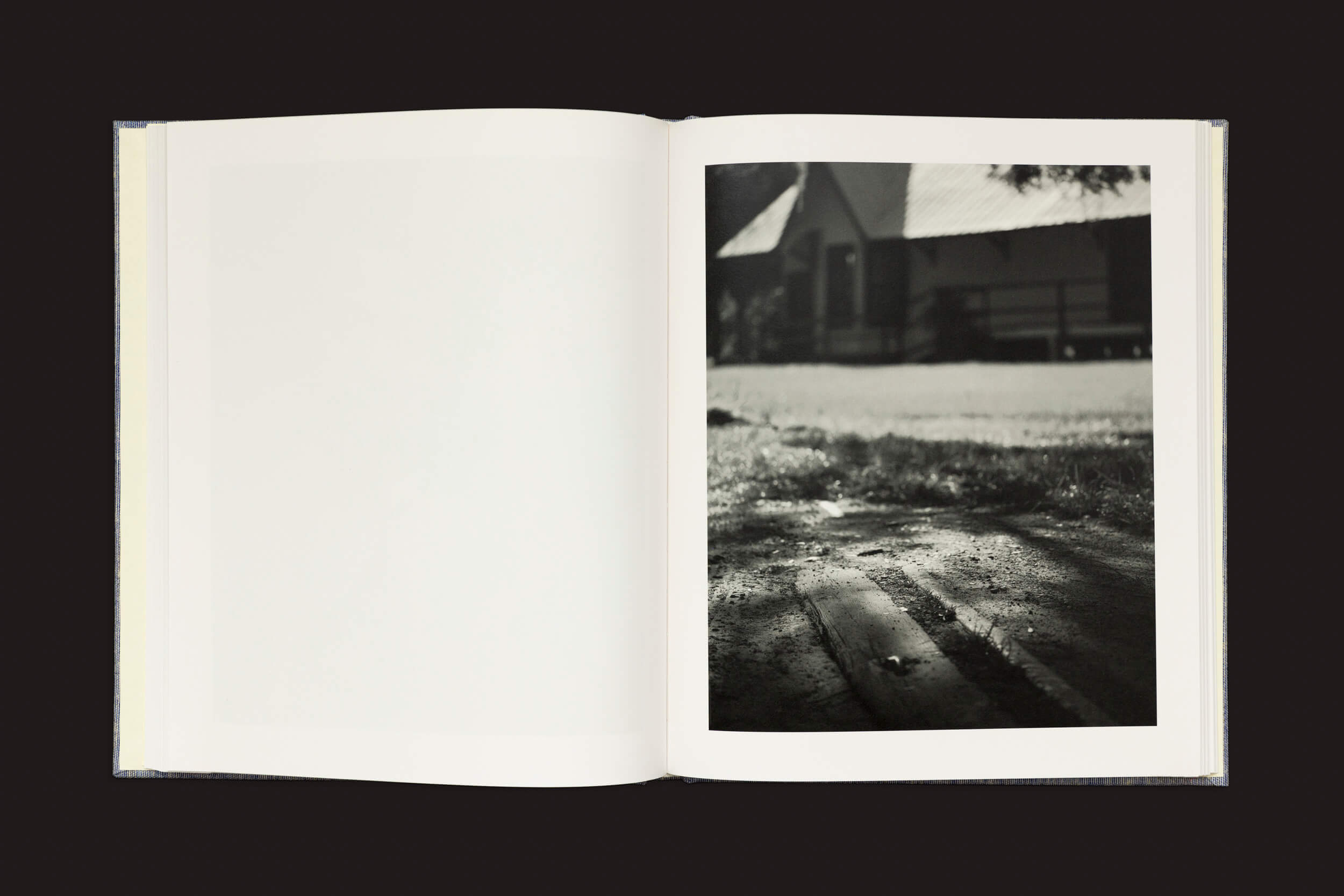 Em uma superfície escura repousa Rahim Fortune: Hardtack by Loose Joints, aberto em uma página mostrando uma foto em preto e branco de uma casa com um telhado inclinado. Um caminho, sombreado por árvores, leva até ela. A página esquerda em branco parece pronta para histórias de tradições culturais.