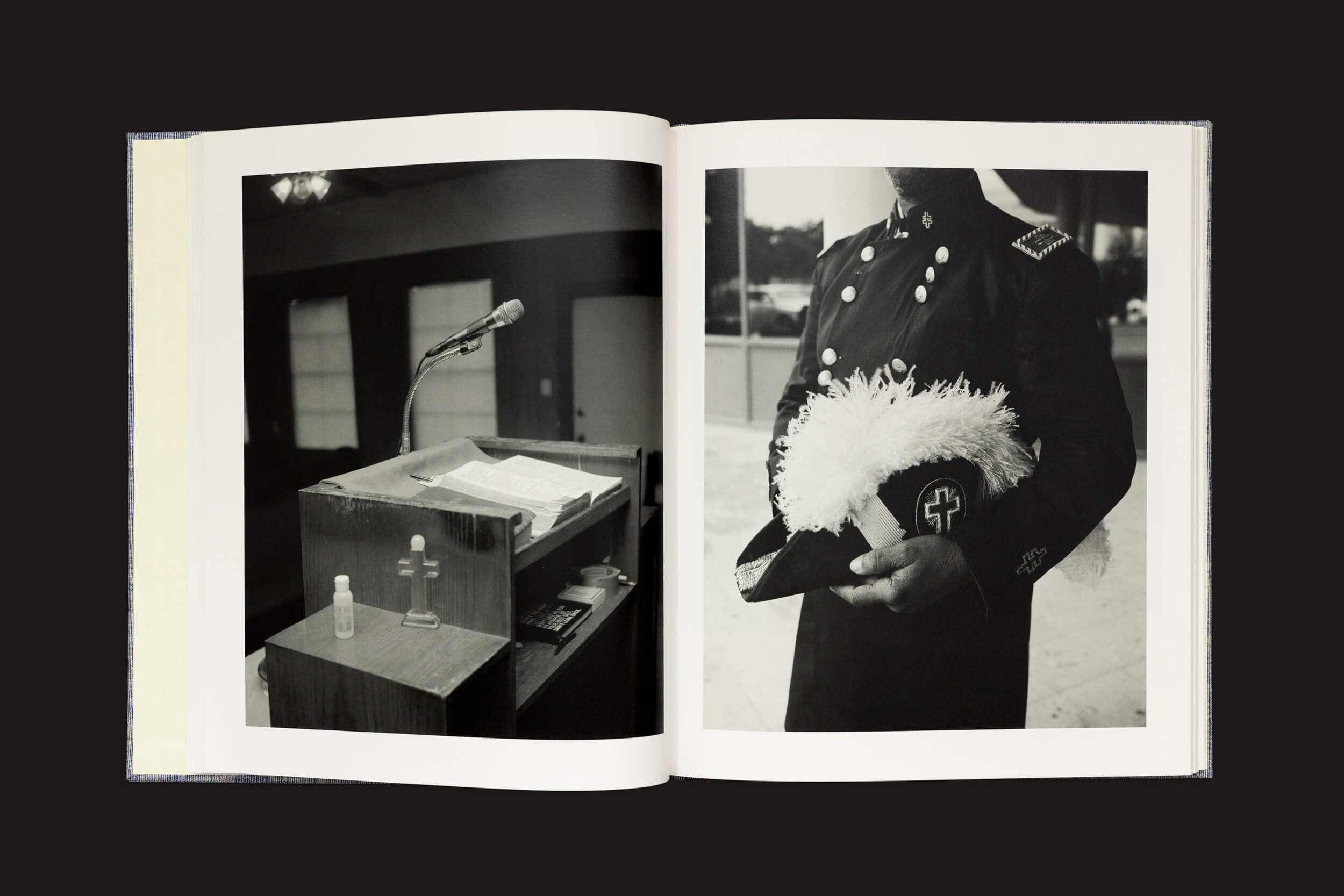 Uma foto em preto e branco intitulada Hardtack por Rahim Fortune, da Loose Joints, captura um livro aberto. À esquerda, um pódio com um microfone, livros e álcool em gel simboliza a América pós-emancipação. À direita, uma pessoa uniformizada com um chapéu enfeitado com penas e uma cruz, refletindo ricas tradições culturais.