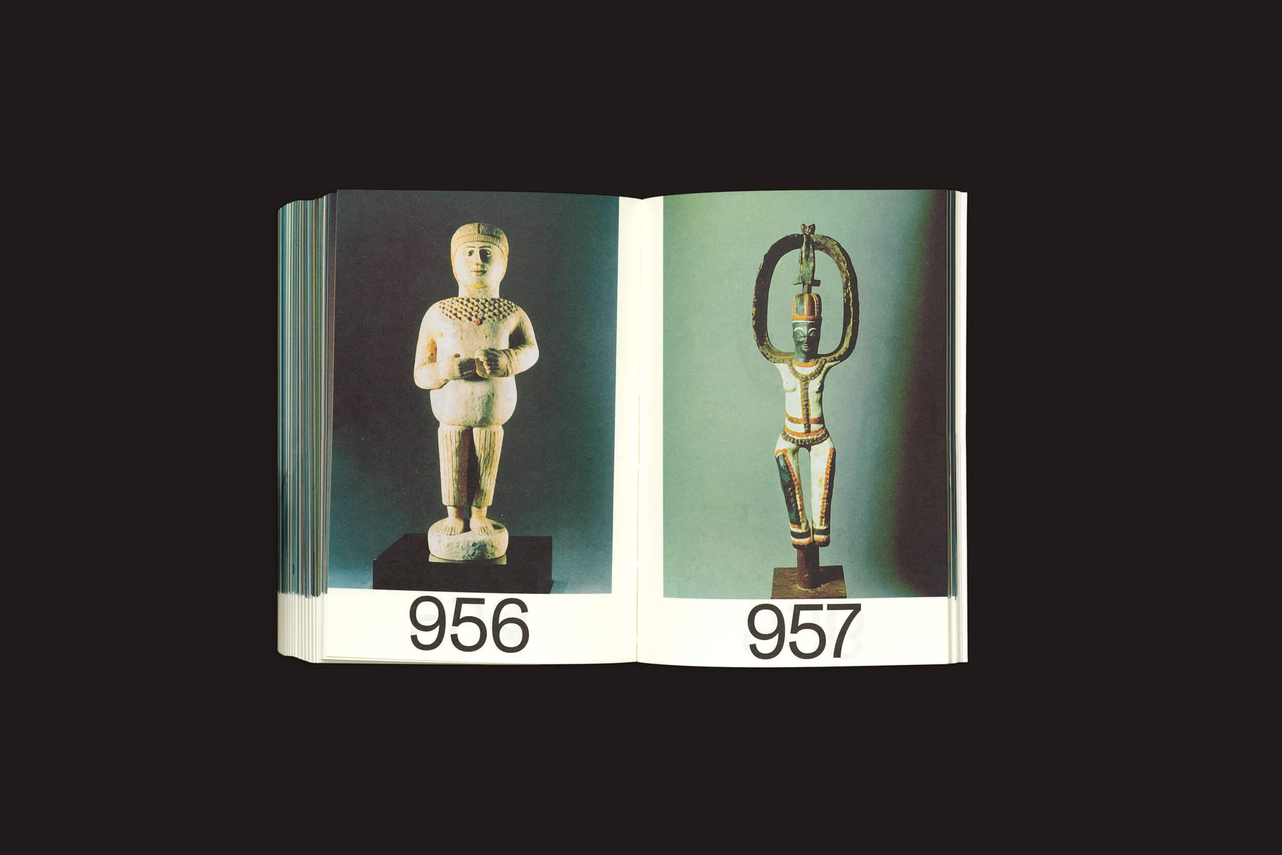 Um livro aberto de Martín Bollati: Hermes/Unesco da SED Editorial exibe imagens de duas esculturas que lembram civilizações antigas. À esquerda, uma pequena estatueta branca etiquetada como 956 e, à direita, uma alta escultura metálica com braços levantados etiquetada como 957, ambas colocadas contra um fundo escuro.