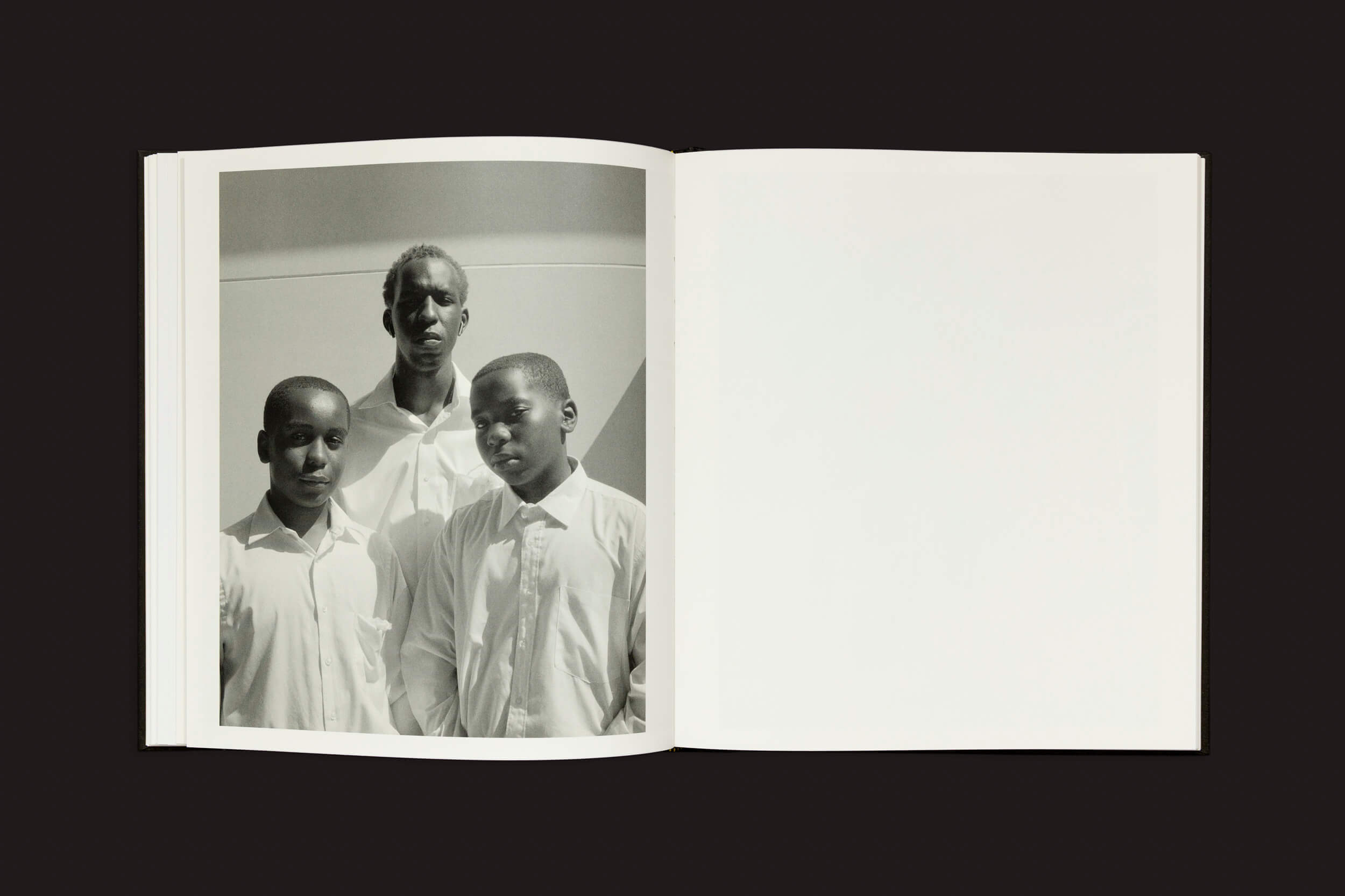 O livro aberto, Rahim Fortune: I cant stand to see you cry de Loose Joints, revela uma fotografia em preto e branco na página esquerda mostrando três pessoas em camisas brancas, capturando identidade compartilhada. A página direita permanece em branco, colocada contra um fundo simples.