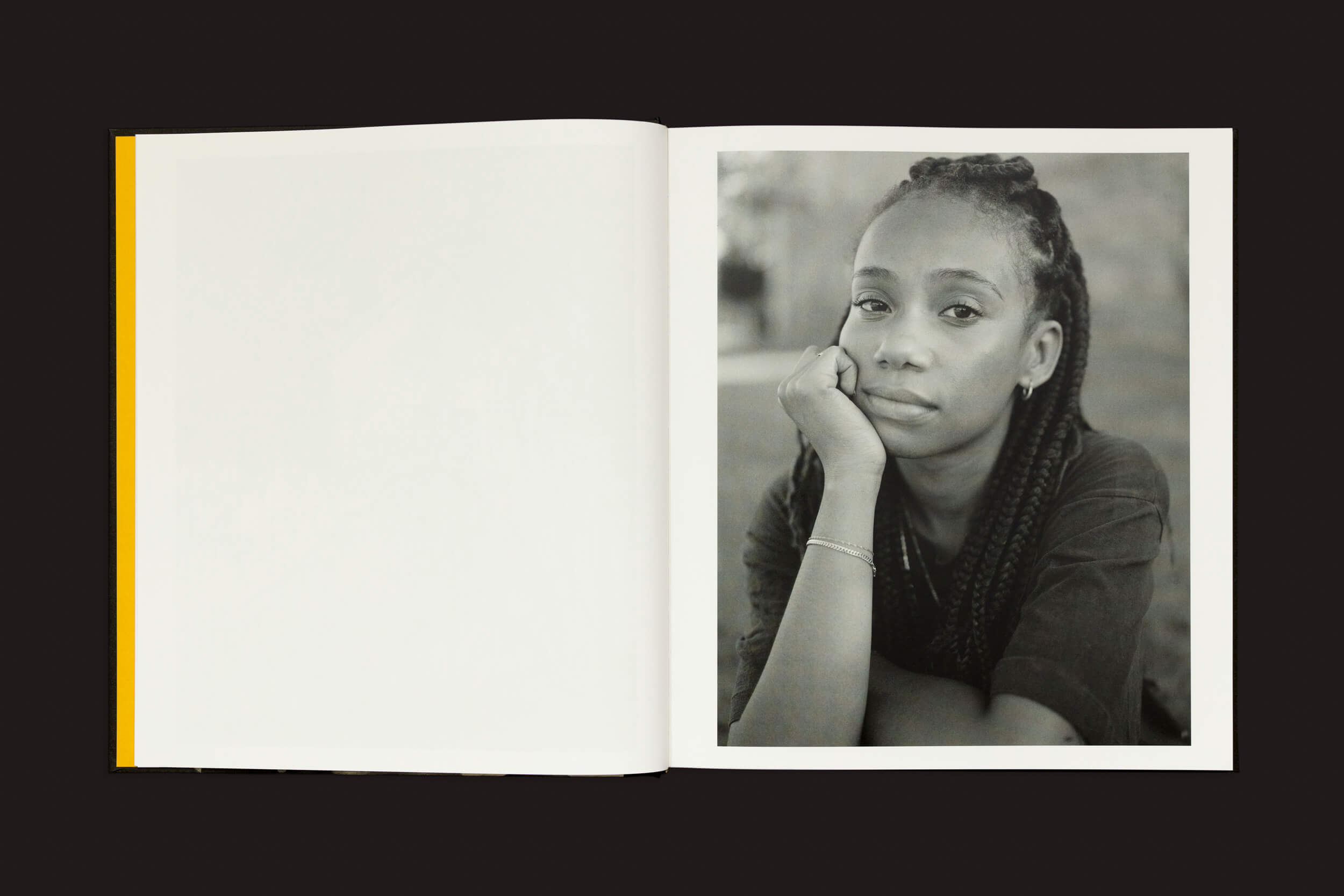 O livro Rahim Fortune: I cant stand to see you cry de Loose Joints está aberto contra um fundo preto, exibindo um retrato cativante de uma pessoa com cabelo trançado apoiando o rosto na mão, transmitindo identidade. A página da esquerda está em branco com uma borda amarela.