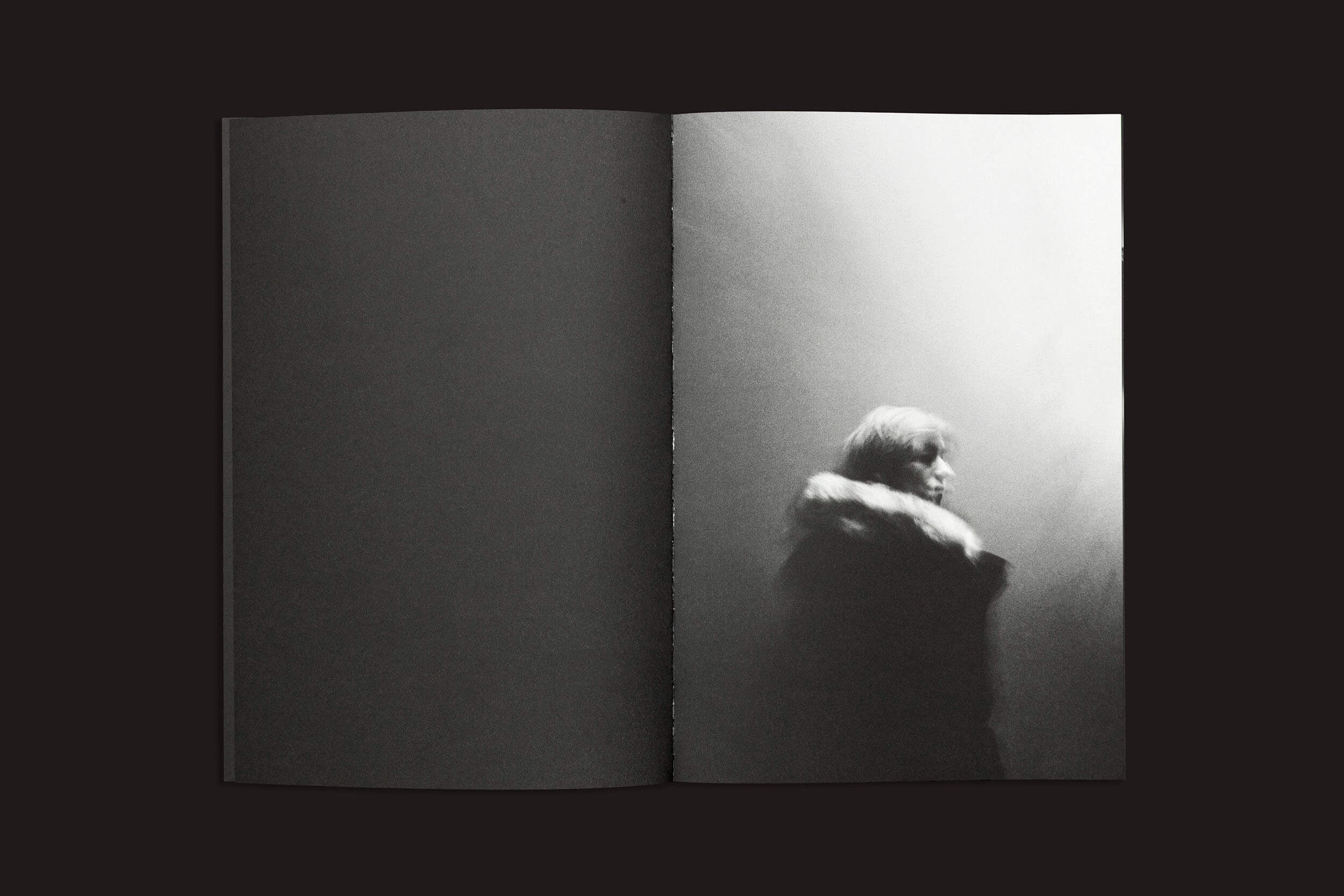 Na imagem em preto e branco de Gui Marcondes, "Eu sei que existo porque você me imagina", da Nearest Truth Editions, um livro aberto mostra uma pessoa com um casaco de gola de pele olhando para a direita. A iluminação suave adiciona tons sombrios, capturando a experiência humana da incerteza contra um fundo indefinido.