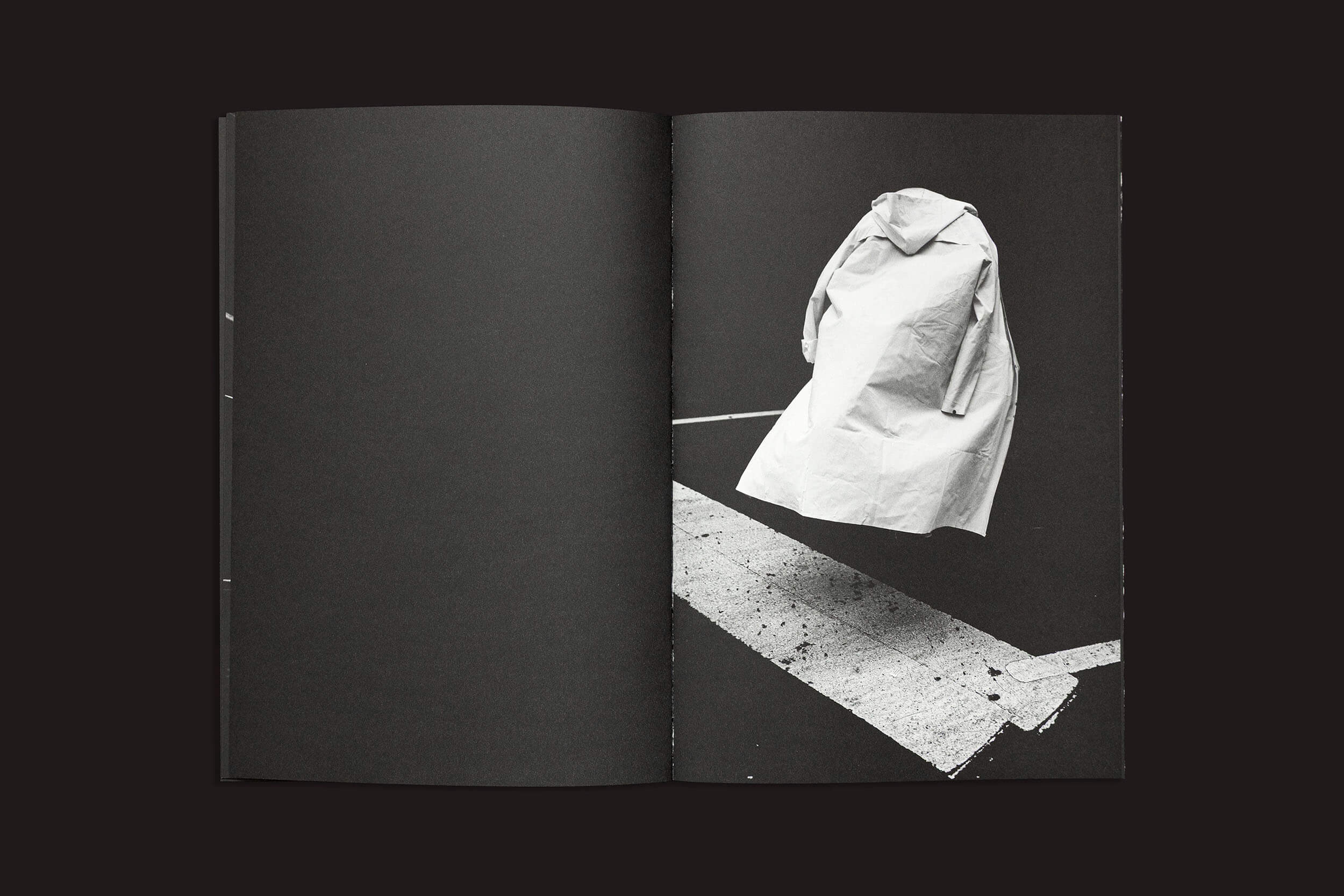 A imagem em preto e branco de "Gui Marcondes: Eu Sei Que Existo Porque Você Me Imagina", da Nearest Truth Editions, apresenta um sobretudo suspenso sobre uma faixa de pedestres. Ela captura a essência surreal da exploração pessoal, com um fundo escuro e uma sombra tênue que evoca a incerteza humana.