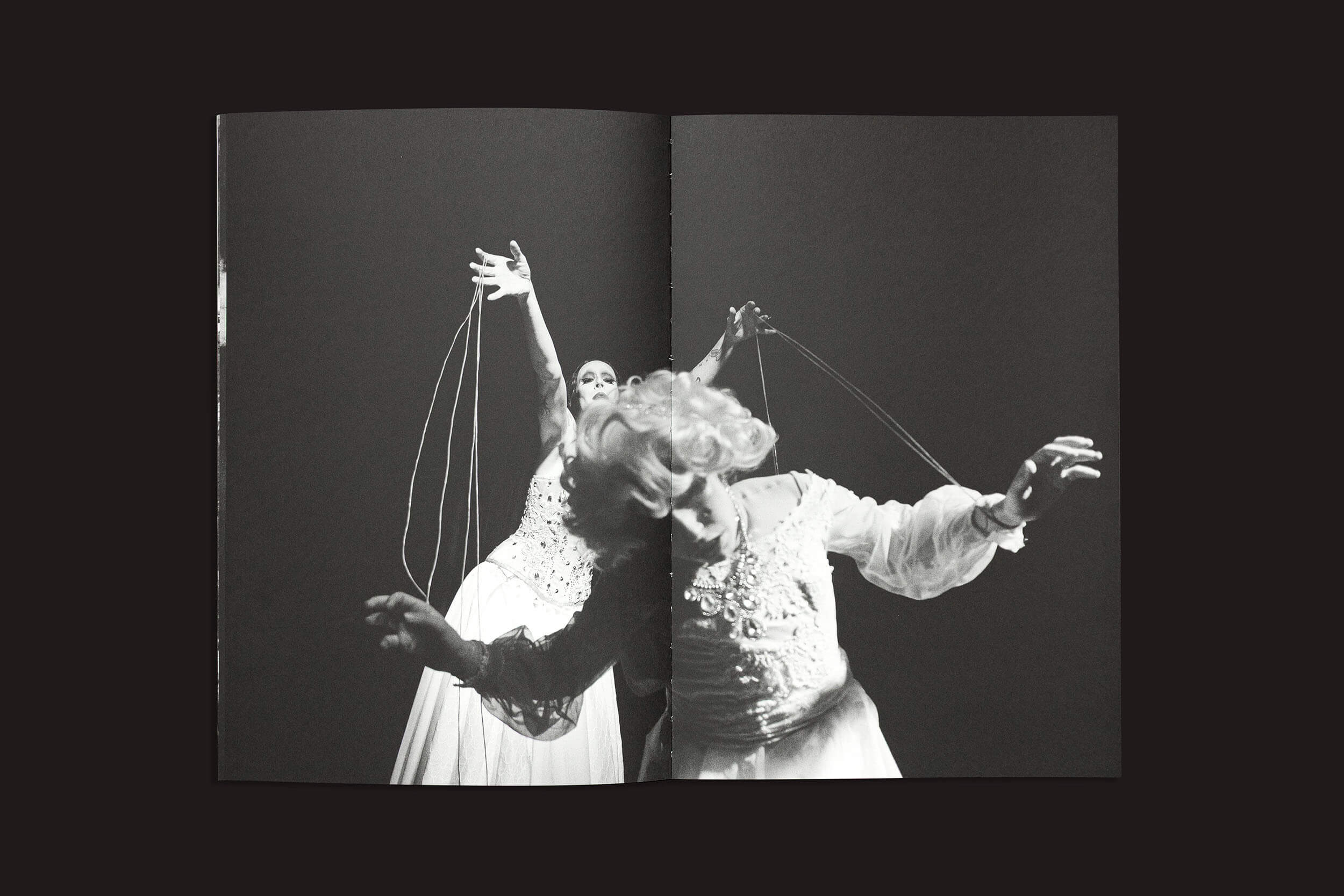 Em um palco escuro, dois artistas encarnam a experiência humana da incerteza de "Gui Marcondes: Eu sei que existo porque você me imagina", da Nearest Truth Editions. Vestidos com figurinos teatrais, um segura cordas no alto enquanto o outro se inclina para a frente, ambos encenando uma cena dramática e expressiva.