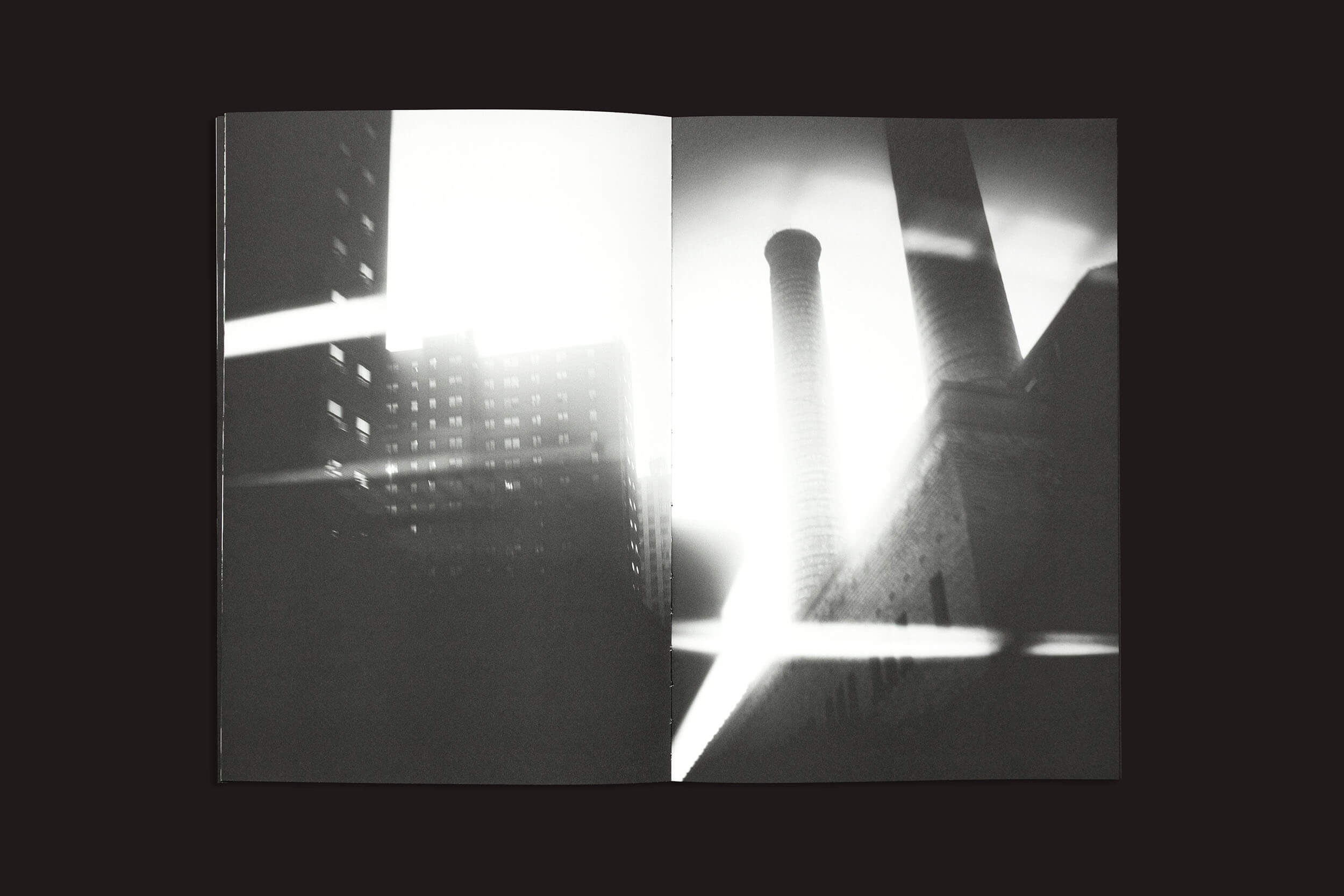 Uma imagem em preto e branco de "Gui Marcondes: Eu sei que existo porque você me imagina", da Nearest Truth Editions, mostra um livro aberto com uma cena industrial de um prédio e duas chaminés altas. Uma luz forte cria um reflexo de lente, refletindo a experiência humana da incerteza, enquanto um arranha-céu é parcialmente visível à esquerda.