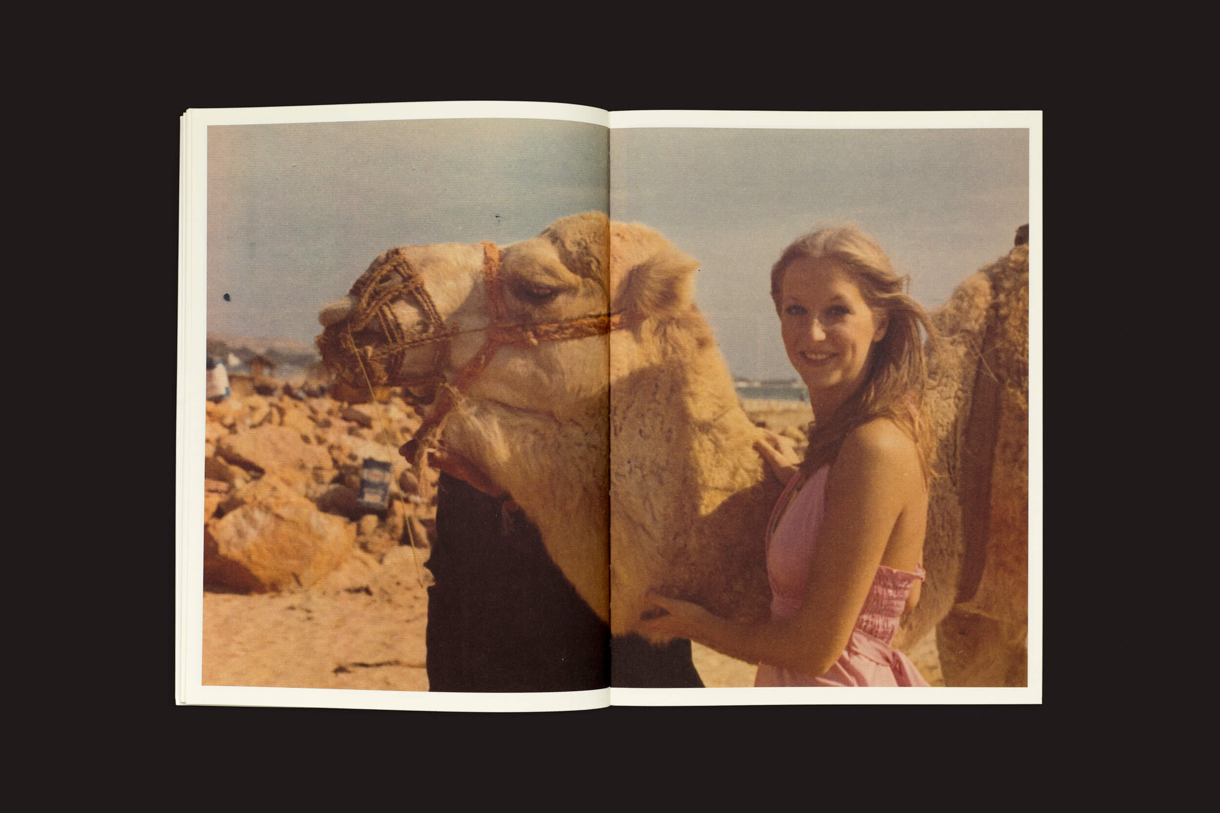 Uma pessoa em um vestido rosa sorri ao lado de um camelo no deserto, contra um fundo rochoso e céu limpo, capturando imagens universais. Esta cena é apresentada em "Incomplete Encyclopedia of Touch" de Erik Kessels, Thomas Sauvin e Karel De Mulder da RVB Books, evocando memórias de álbuns de família.