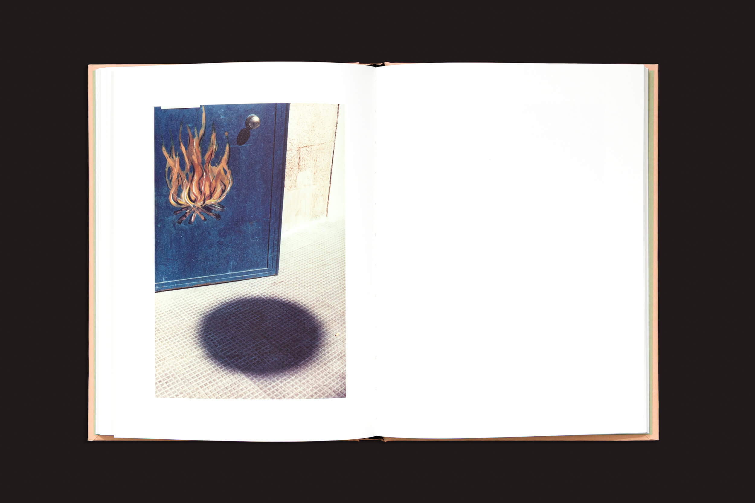 Sobre uma superfície escura, um exemplar aberto de Federico Clavarino: Italia O Italia, da Void, revela a imagem de uma porta azul com um motivo de chamas, projetando uma sombra circular no piso texturizado. As páginas, que lembram a fotografia simbólica de Clavarino, permanecem em branco.