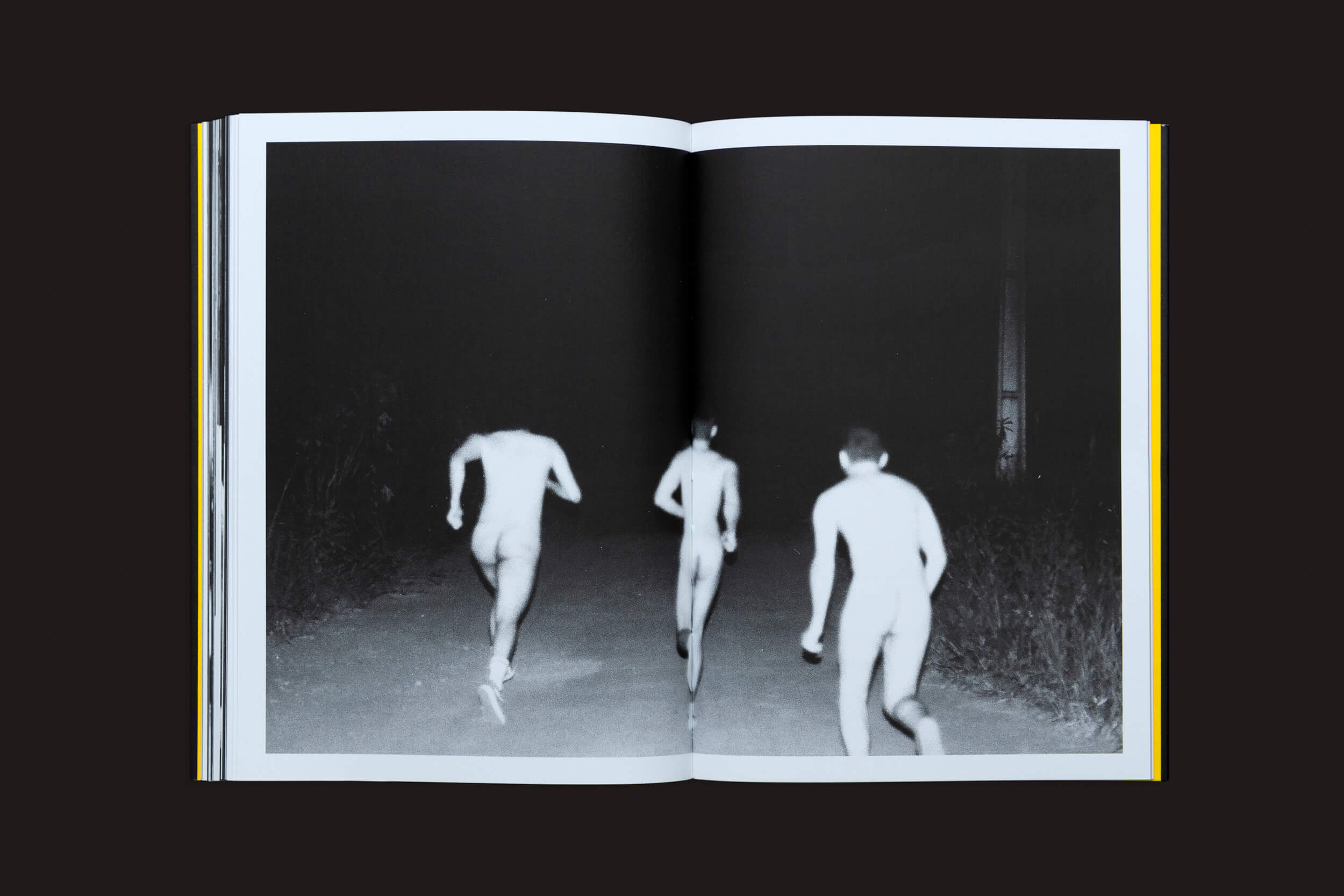 Um livro aberto, Artur Lahoz: Klexos de Selo Turvo, apresenta uma foto em preto e branco de três indivíduos nus correndo em um caminho. Capturados de trás, dois estão à esquerda e um à direita, simbolizando a natureza transitória da memoria.